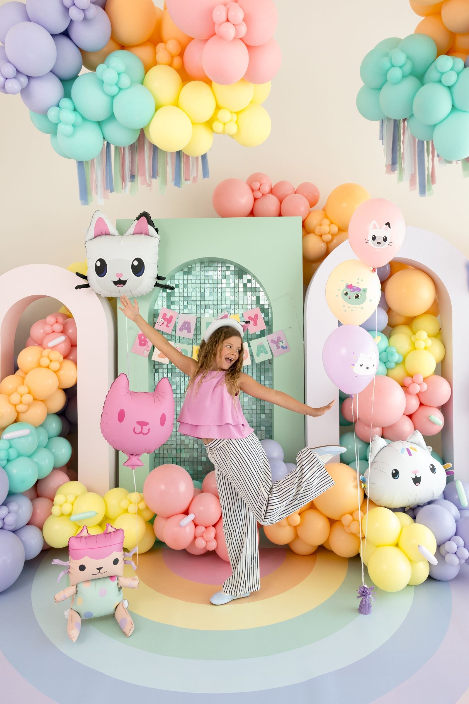 Gabby's Dollhouse themed birthday party decorations featuring character foil balloons, a Happy Birthday banner, and a purple striped tablecloth.