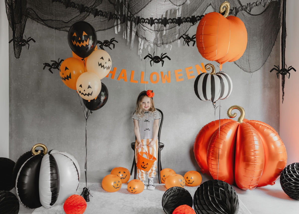 A child standing in a Halloween-decorated room. Large orange and black and white striped pumpkin foil balloons are visible, latex balloons with jack-o'-lantern faces, and spider decorations.