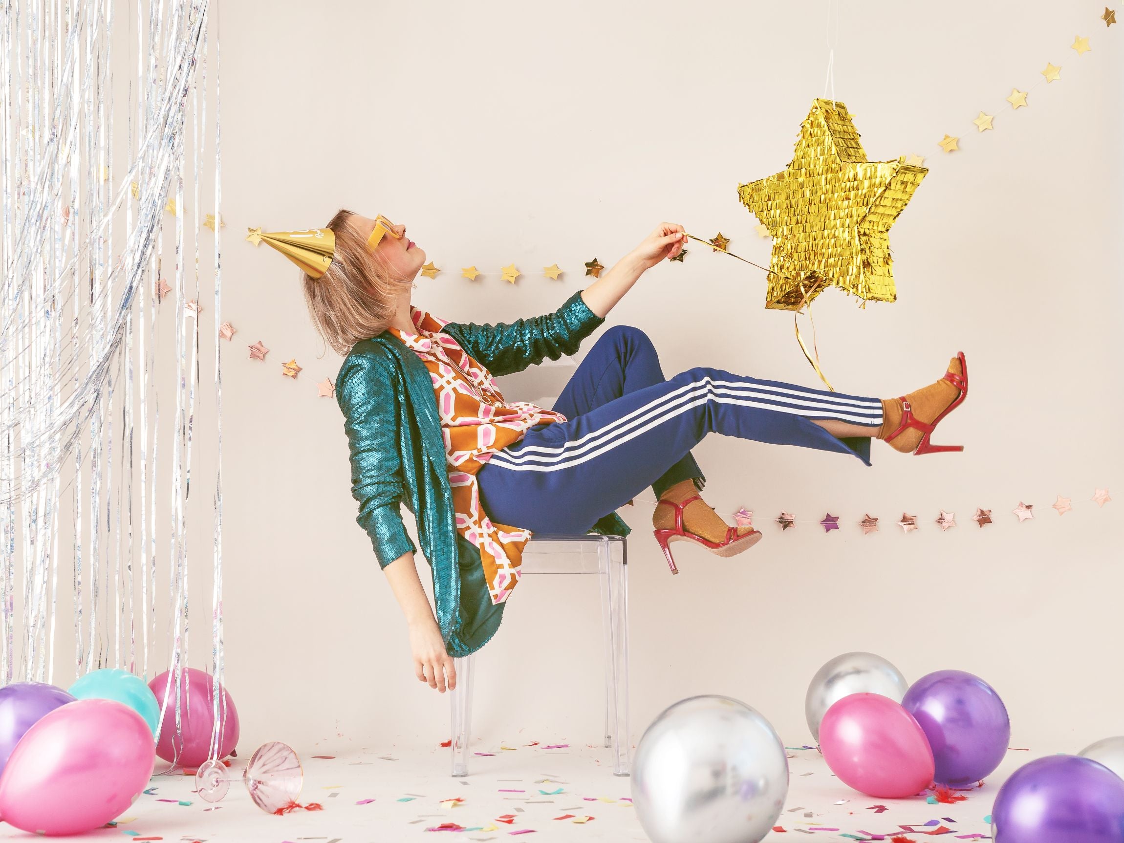 A person wearing a gold party hat and sunglasses, dressed in a green sequin jacket, colorful patterned shirt, and blue striped pants, appearing to float or lean back dramatically while reaching towards a gold star-shaped piñata, in a festive party setting with balloons and streamers.