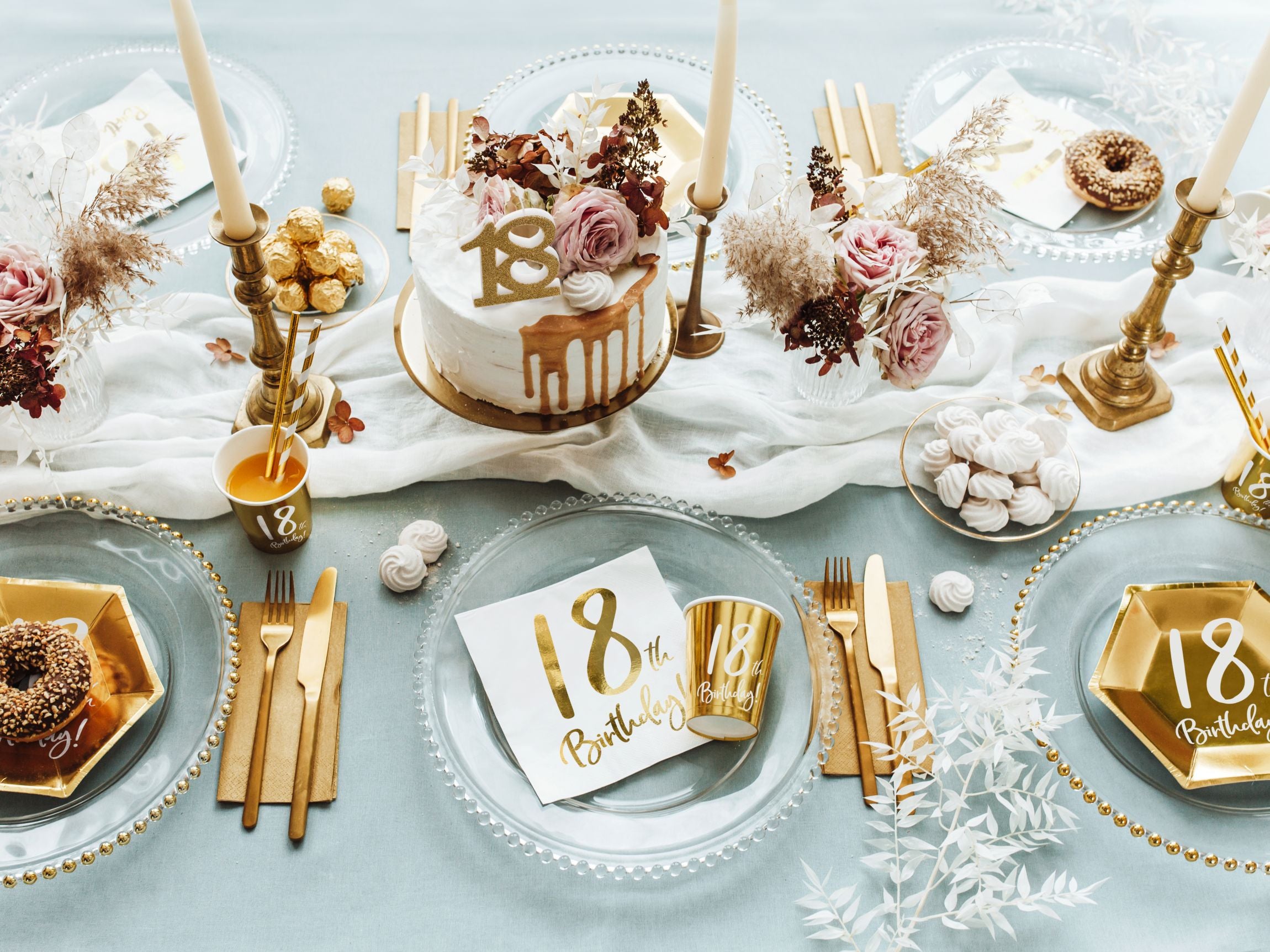 An overhead view of a richly decorated table for an 18th birthday party. The table features white paper napkins with gold "18th Birthday!" print, golden hexagonal plates, a white birthday cake with gold drip, tall white candles in gold holders, and floral arrangements, all on a light blue tablecloth.