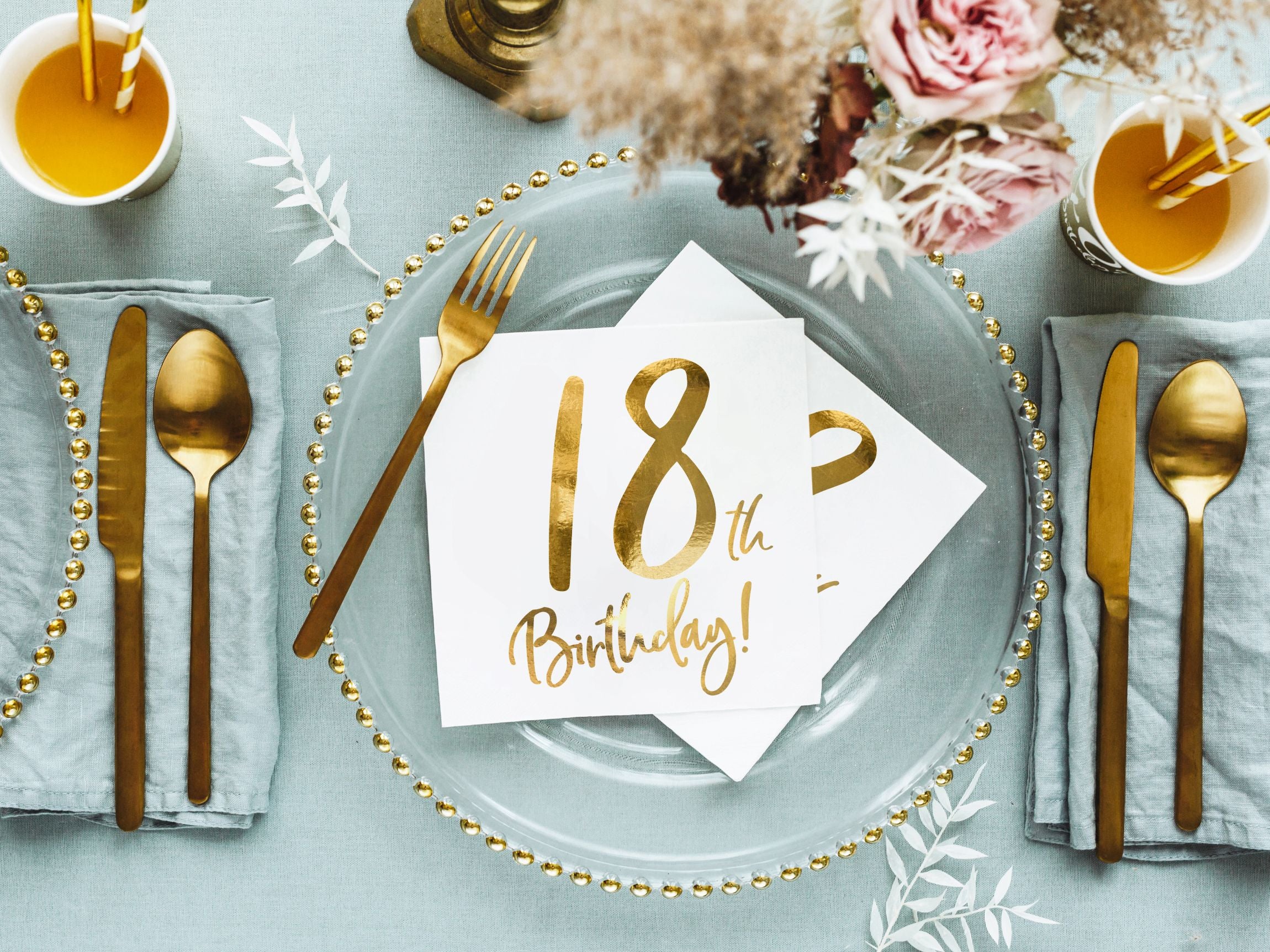 A close-up overhead view of a single place setting on an 18th birthday party table. A clear glass charger plate sits on a light blue napkin, topped with a white paper napkin featuring a gold "18th Birthday!" inscription. Gold cutlery is placed on either side, with a small vase of dried flowers and a drink in the background.