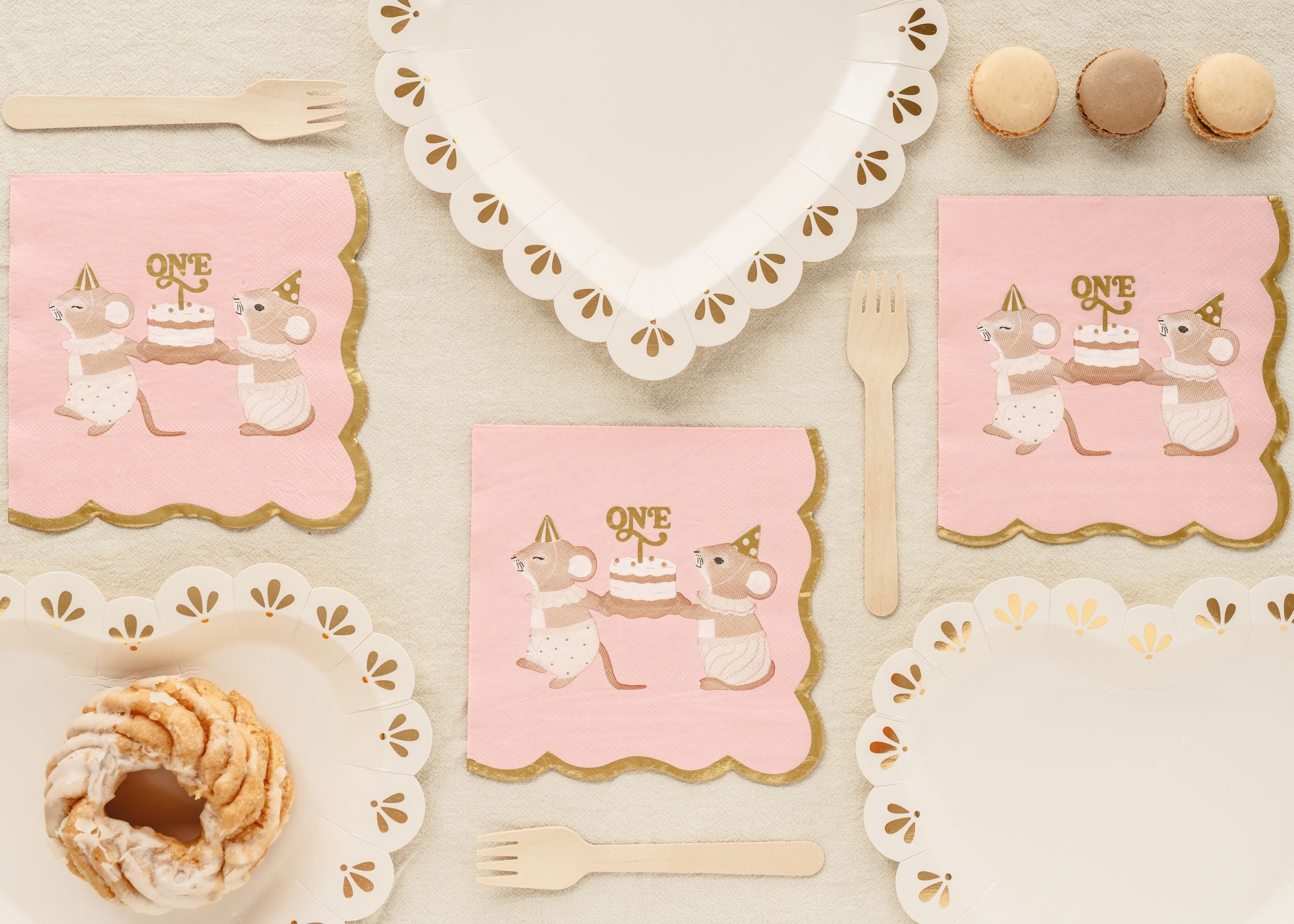 A top-down view of pink 1st birthday napkins with a mice and cake design, styled with white heart-shaped plates, wooden cutlery, and macarons.