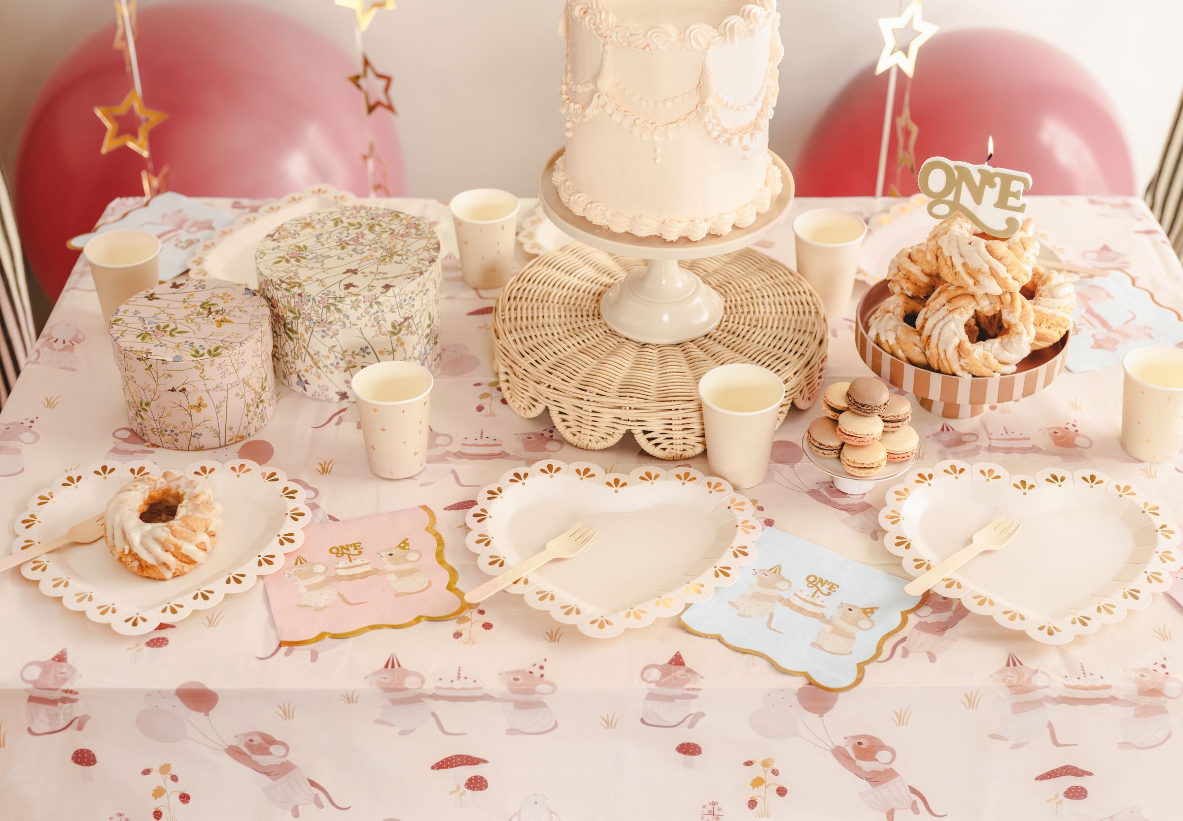 A beautifully styled 1st birthday party table featuring a white cake on a rattan stand, pink balloons, star cups, and pastel pink mice napkins.