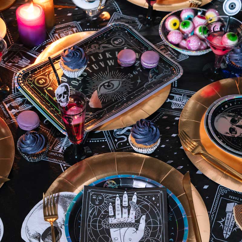 A dark Halloween party table setting featuring gold plates, purple cupcakes, and a black napkin printed with a white esoteric hand and snake design.