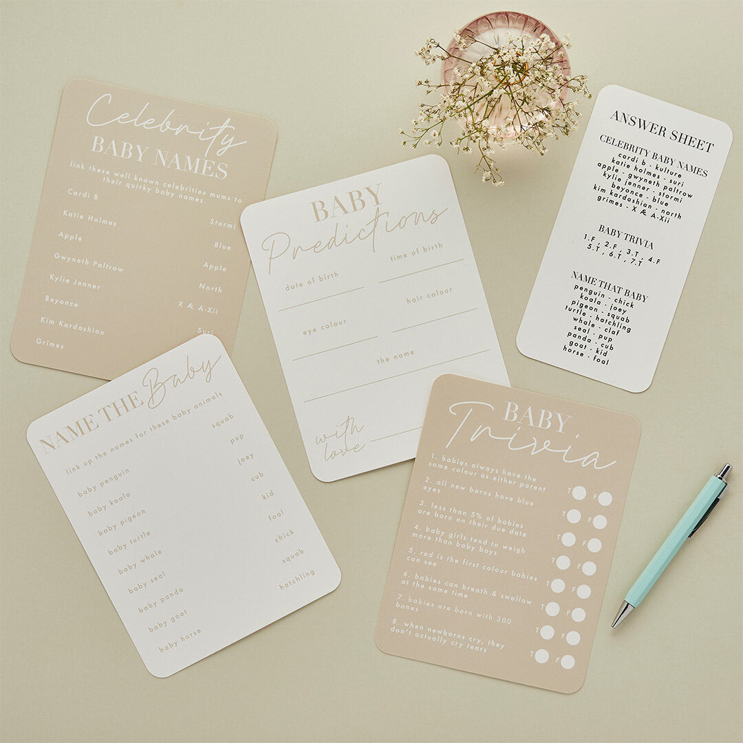 A collection of tan and cream baby shower game cards including Baby Trivia, Predictions, and Name the Baby, laid out with a pen and flowers.
