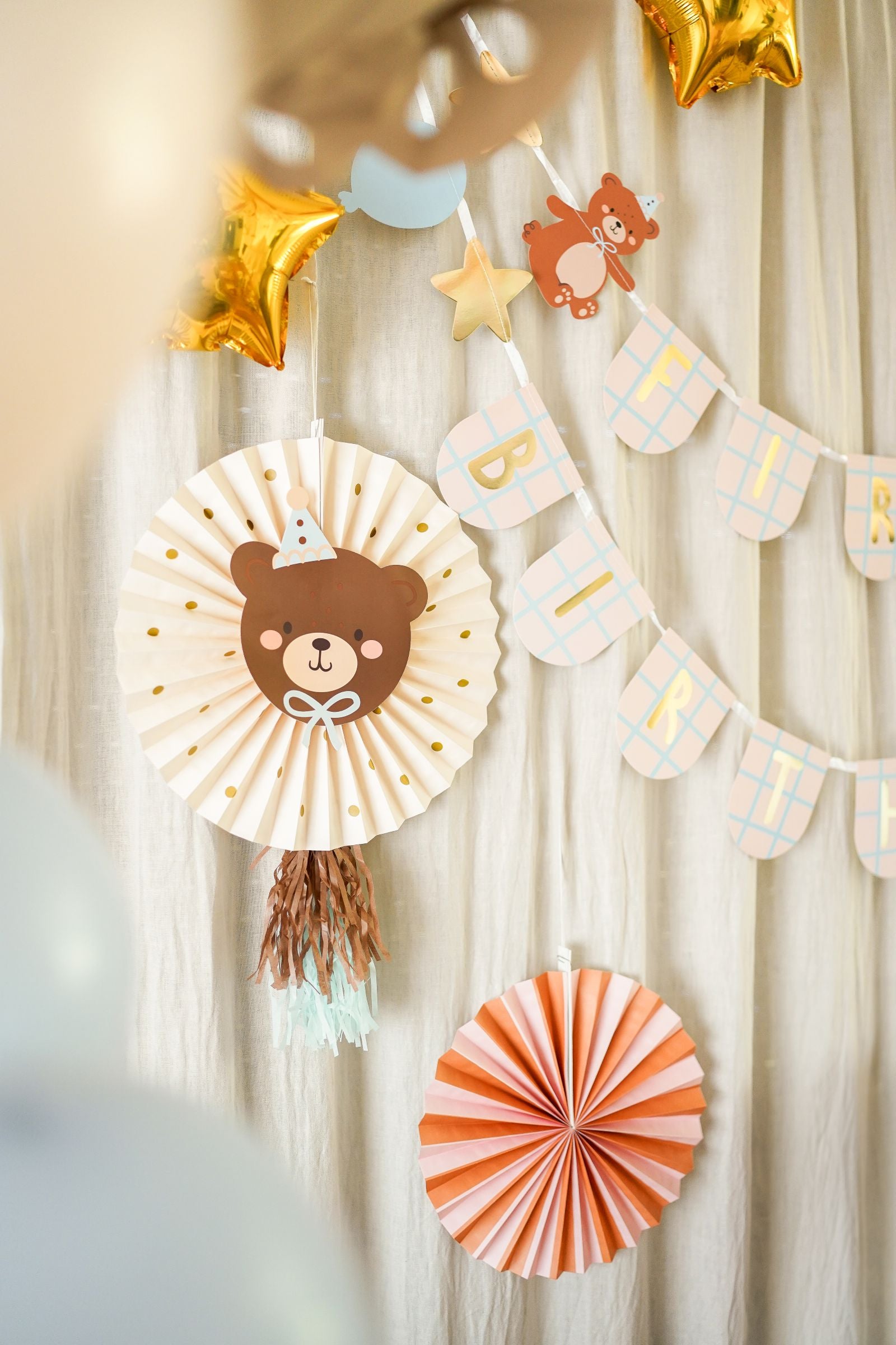 A wide shot of a teddy bear-themed first birthday party setup with a decorated table featuring a cake, balloons, a number "1" balloon, and various party supplies, with a "FIRST BIRTHDAY" banner and a decorative teddy bear rosette hanging in the background against a window with light curtains.