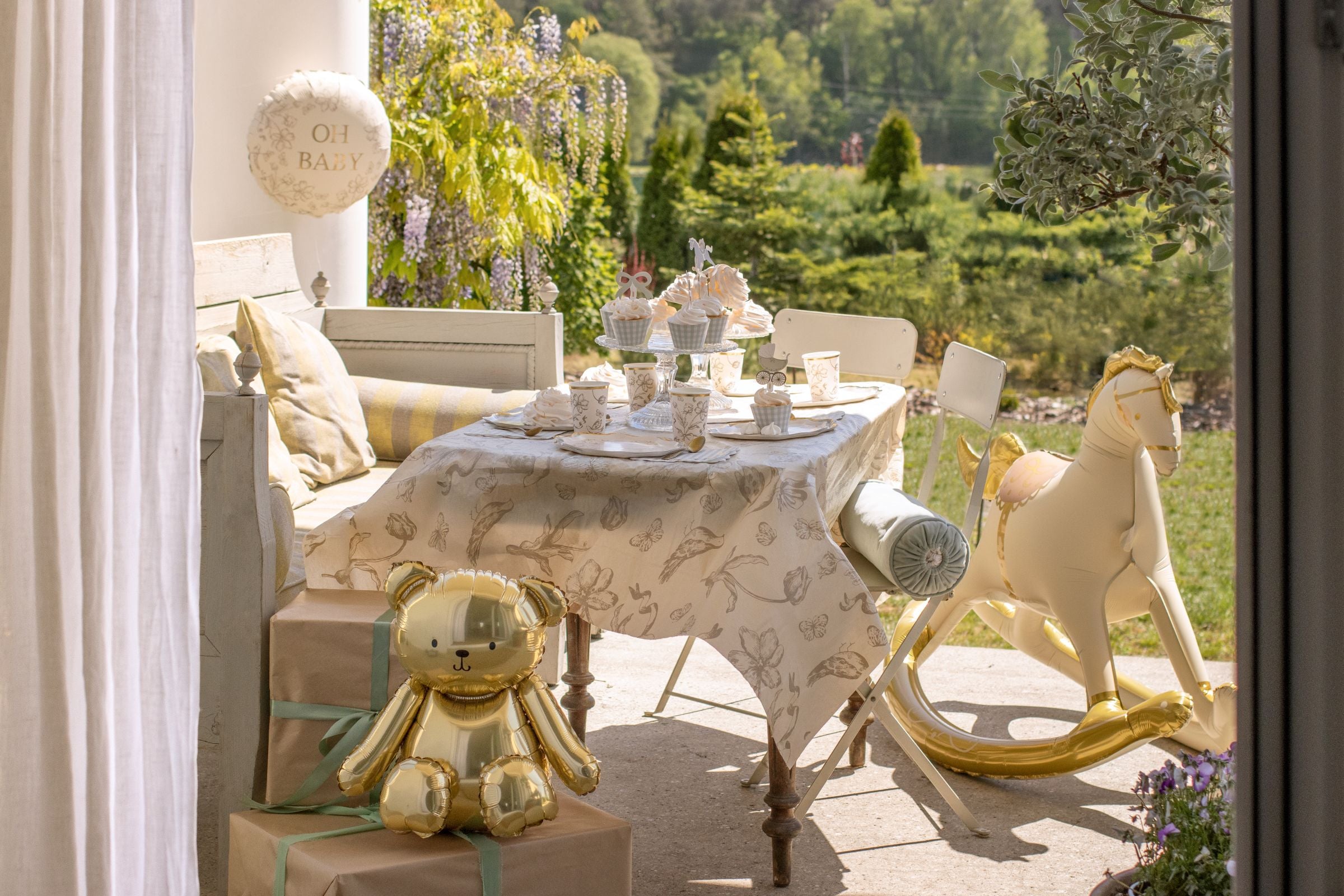 Outdoor party table covered with a beige floral tablecloth next to a gold rocking horse balloon.