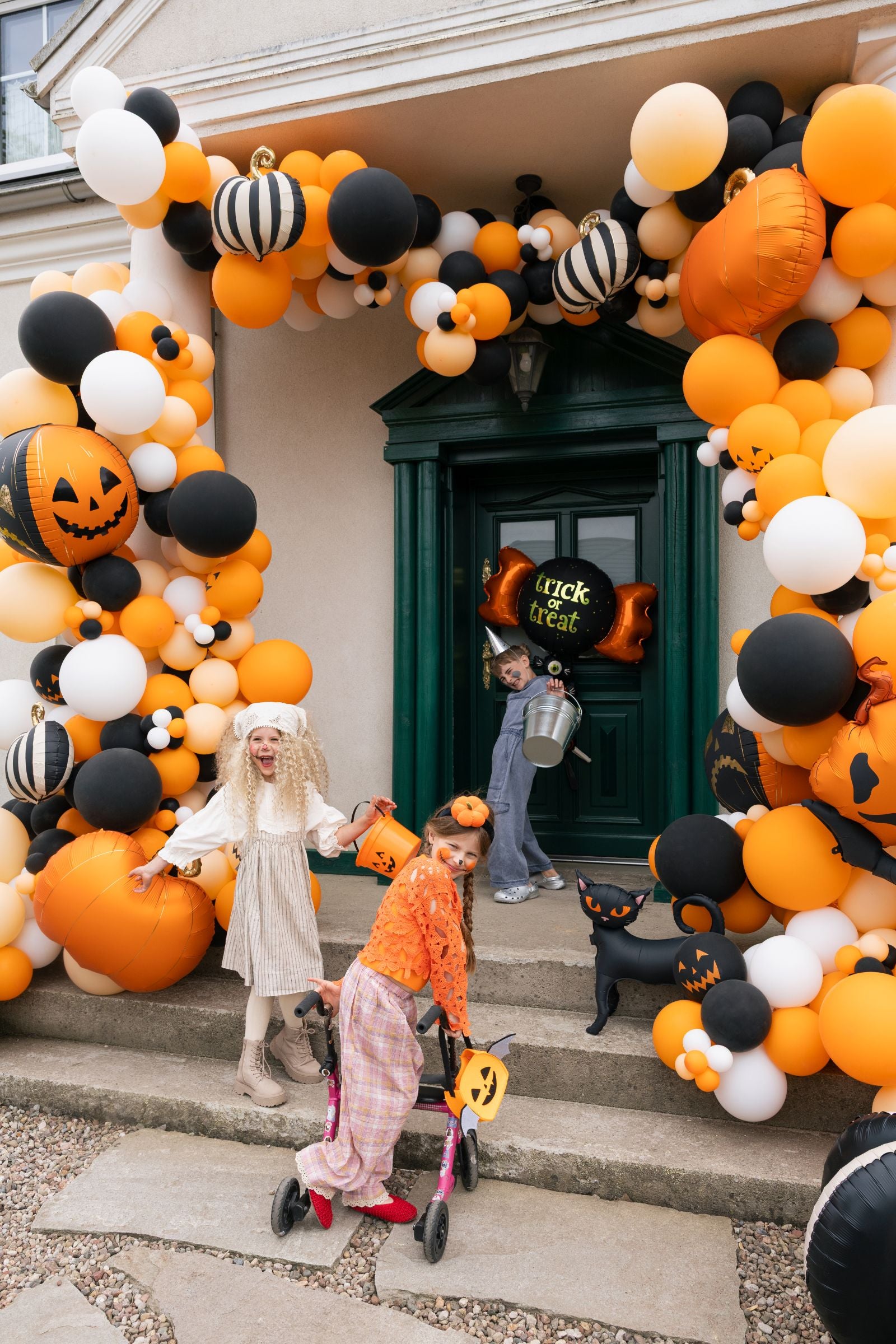 Three children in Halloween costumes stand on the steps of a house decorated with a large orange, black, and white balloon garland featuring pumpkins and jack-o'-lanterns. One child pushes a toy cart, while another holds a bucket and the third wears a ghost costume.
