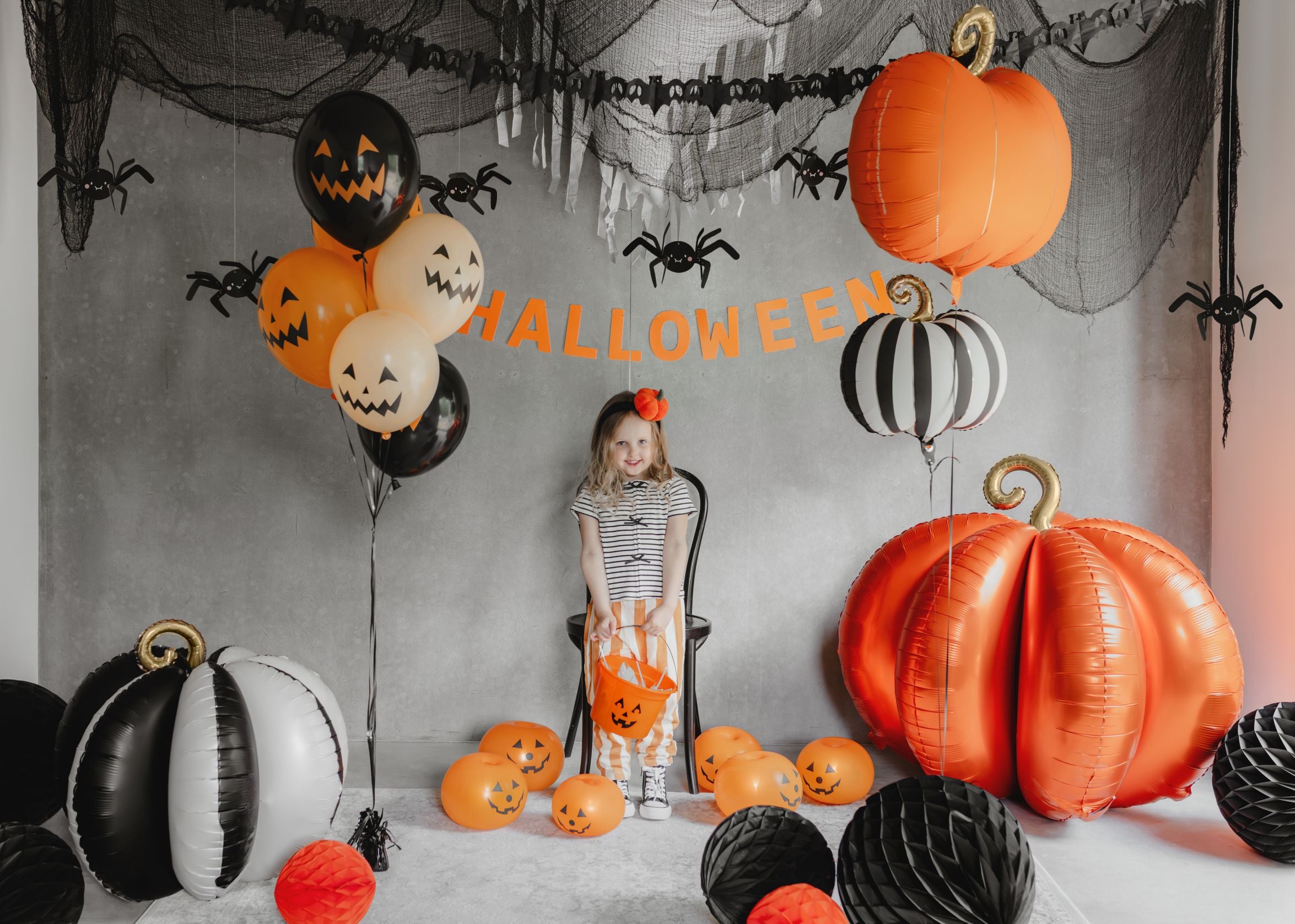 A child in a striped top and orange checkered overalls holding a pumpkin bucket, standing in a Halloween-decorated room. Large orange and black and white striped pumpkin foil balloons are visible, along with black and orange jack-o'-lantern balloons and black honeycomb balls, against a grey wall with cobweb netting and spider decorations.