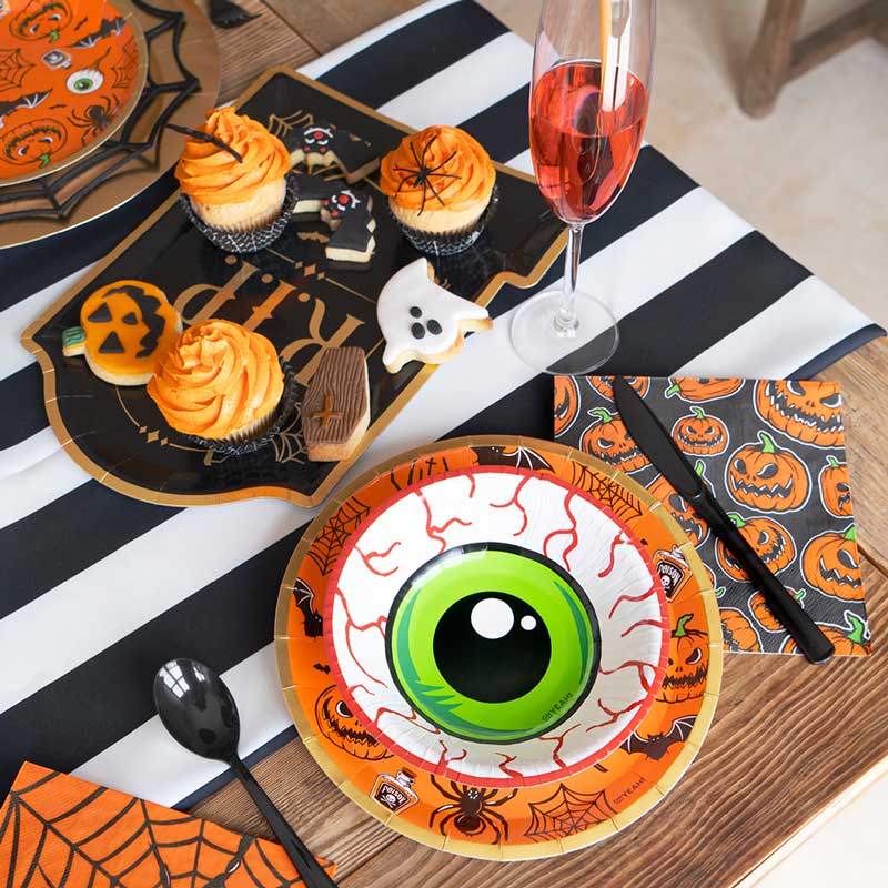 Halloween-themed table setting with orange and black plates, cupcakes, a wine glass, napkins, and spoons arranged on a black-and-white striped tablecloth.