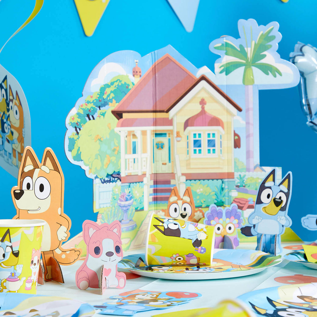 A fully decorated Bluey themed party table showcasing paper cups, plates, and the 7-piece table decoration kit including the Heeler house backdrop and character cut-outs.
