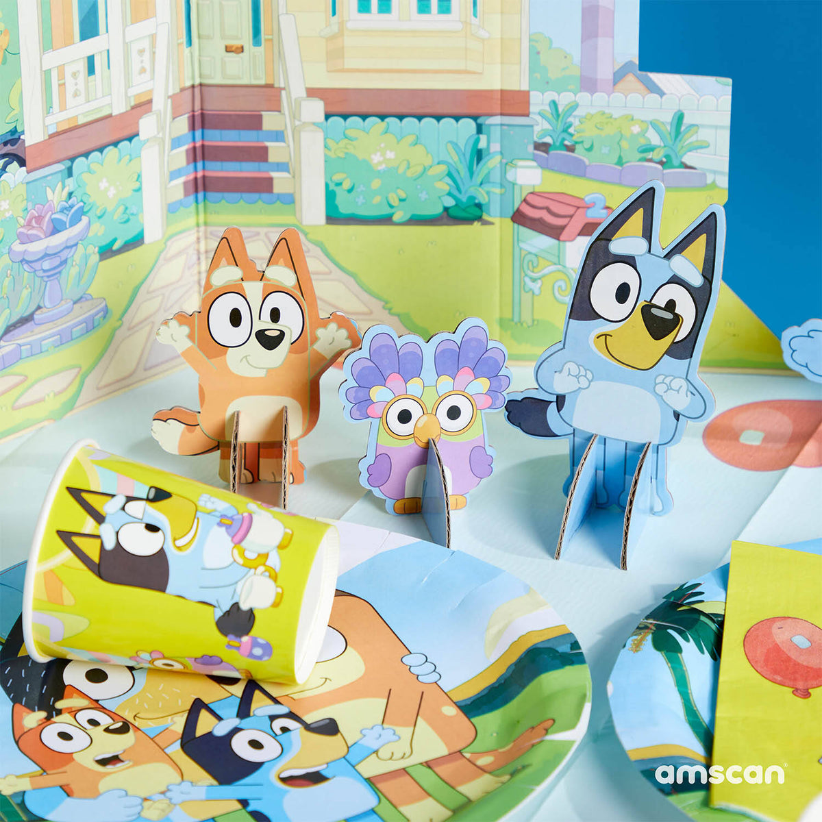 Close-up of Bluey, Bingo, and Chattermax cardboard table decorations standing in front of the printed Heeler house backdrop next to themed party cups.