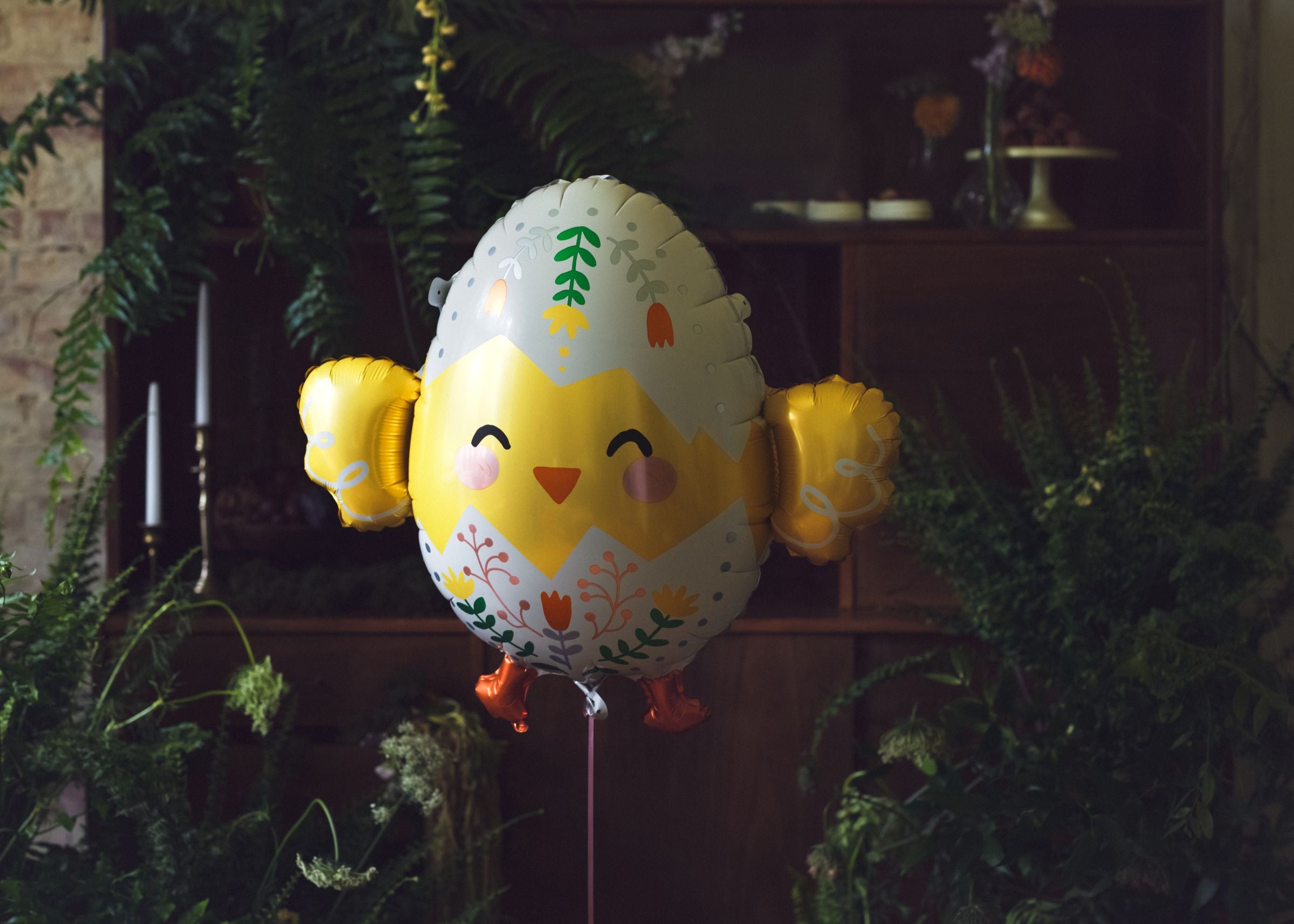 Detailed chick egg foil balloon floating in front of a lush greenery and floral backdrop, highlighting the metallic gold finish.