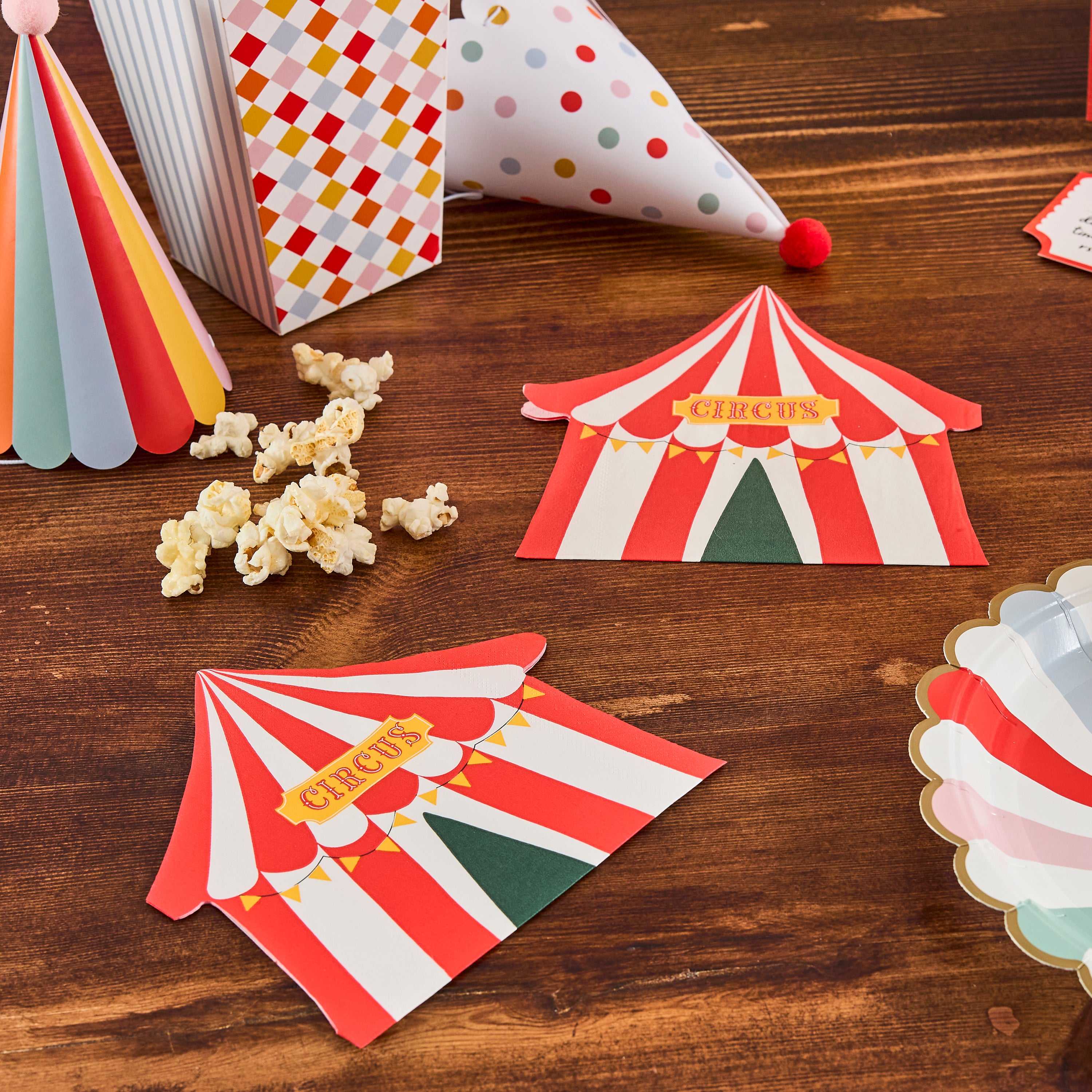 Vintage circus tent napkins displayed on a wooden table next to popcorn boxes, party hats, and striped paper plates.
