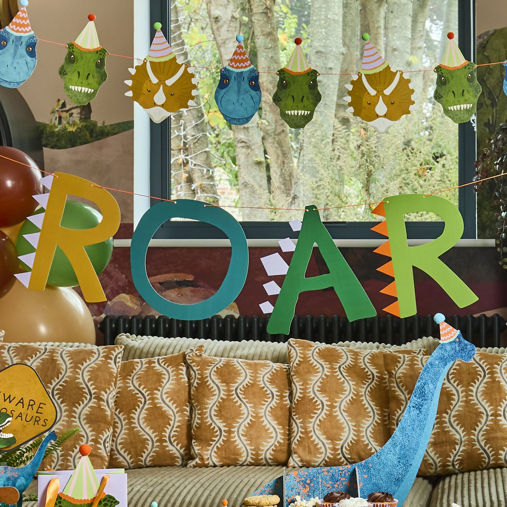 A living room decorated for a dinosaur party with ROAR bunting, dinosaur head garlands, and a blue T-Rex standee.
