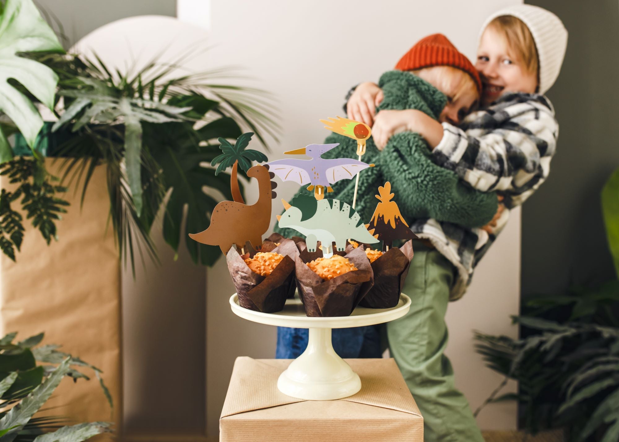 Two children with a dinosaur-themed cupcake stand and decorations indoors.
