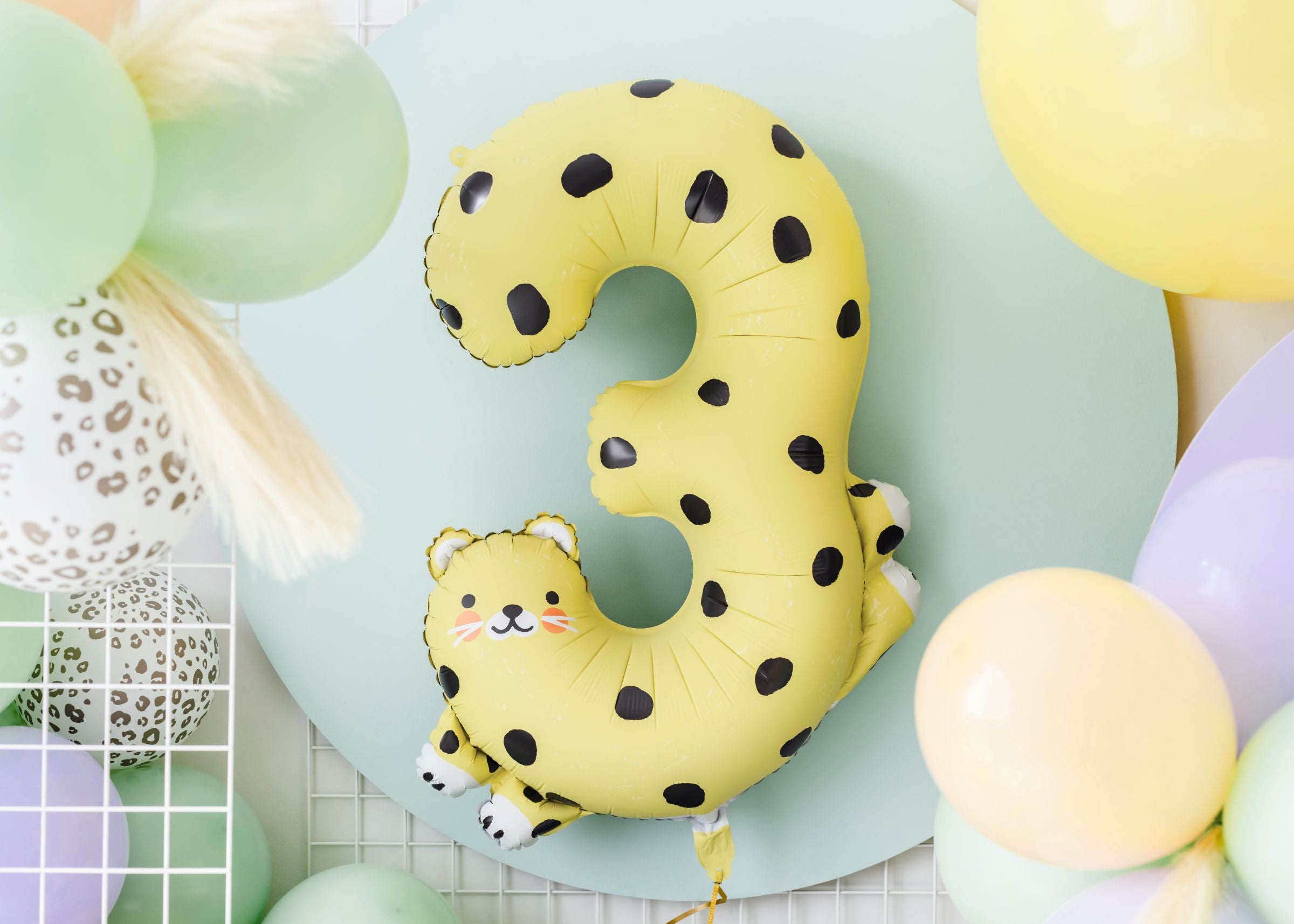 A party scene featuring a large yellow number "3" foil balloon with a leopard print and a smiling cartoon leopard face. It is surrounded by a balloon arch of various pastel-colored balloons, including light peach, green, and lavender, with animal print balloons and beige pampas grass.