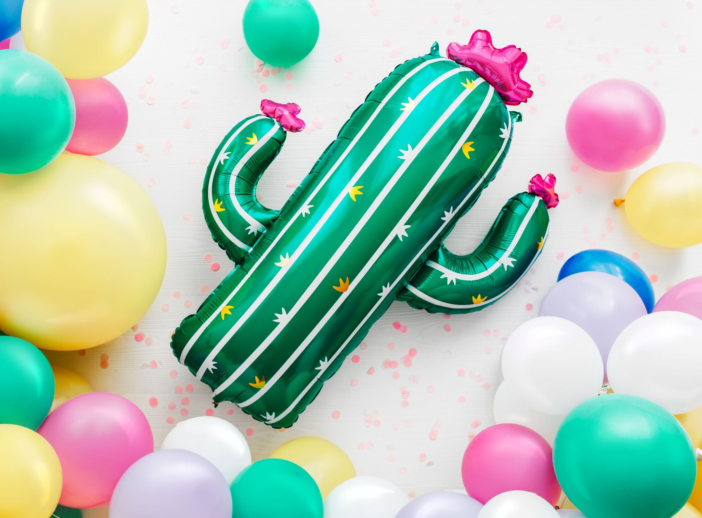 A large, inflated green cactus foil balloon with a pink flower on top, surrounded by a colorful assortment of round latex balloons, including white, yellow, pink, and turquoise, on a textured white background.