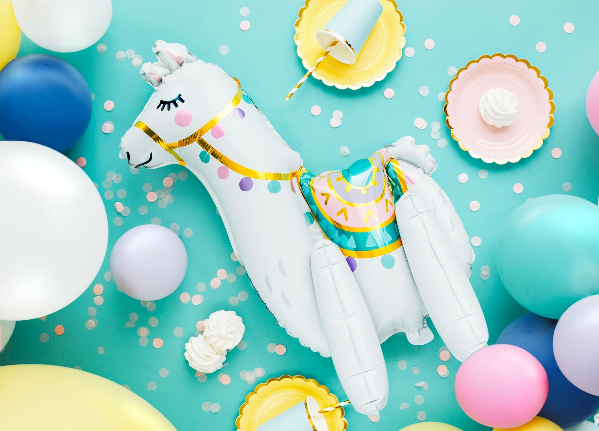 An overhead flat lay of a party scene on a light blue background, featuring a large white llama-shaped foil balloon with colorful printed details. It is surrounded by round latex balloons (white, blue, pink, lavender, and yellow), along with party cups, plates, and confetti.