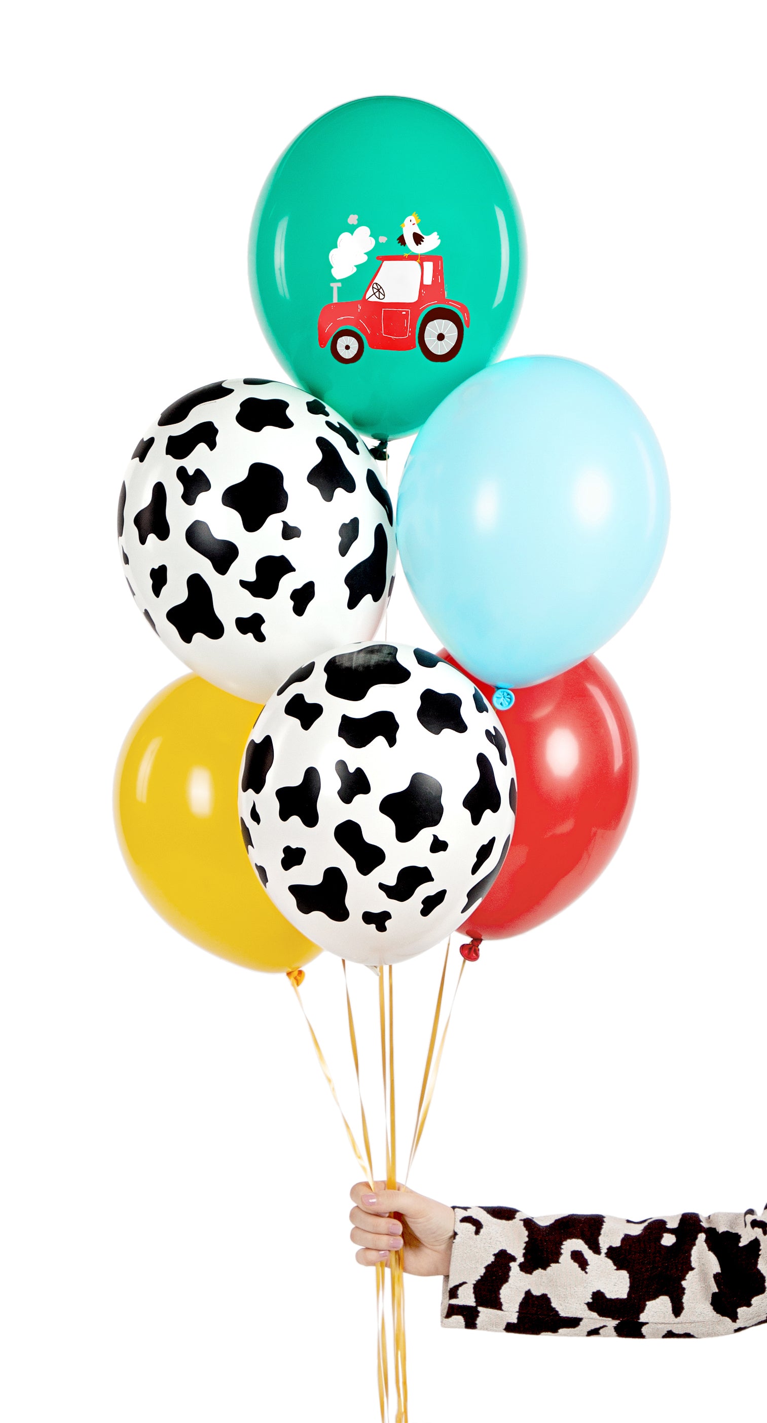 A hand holding a bouquet of six colorful farm-themed balloons with gold ribbons, including cow prints and a tractor design, set against a white background.