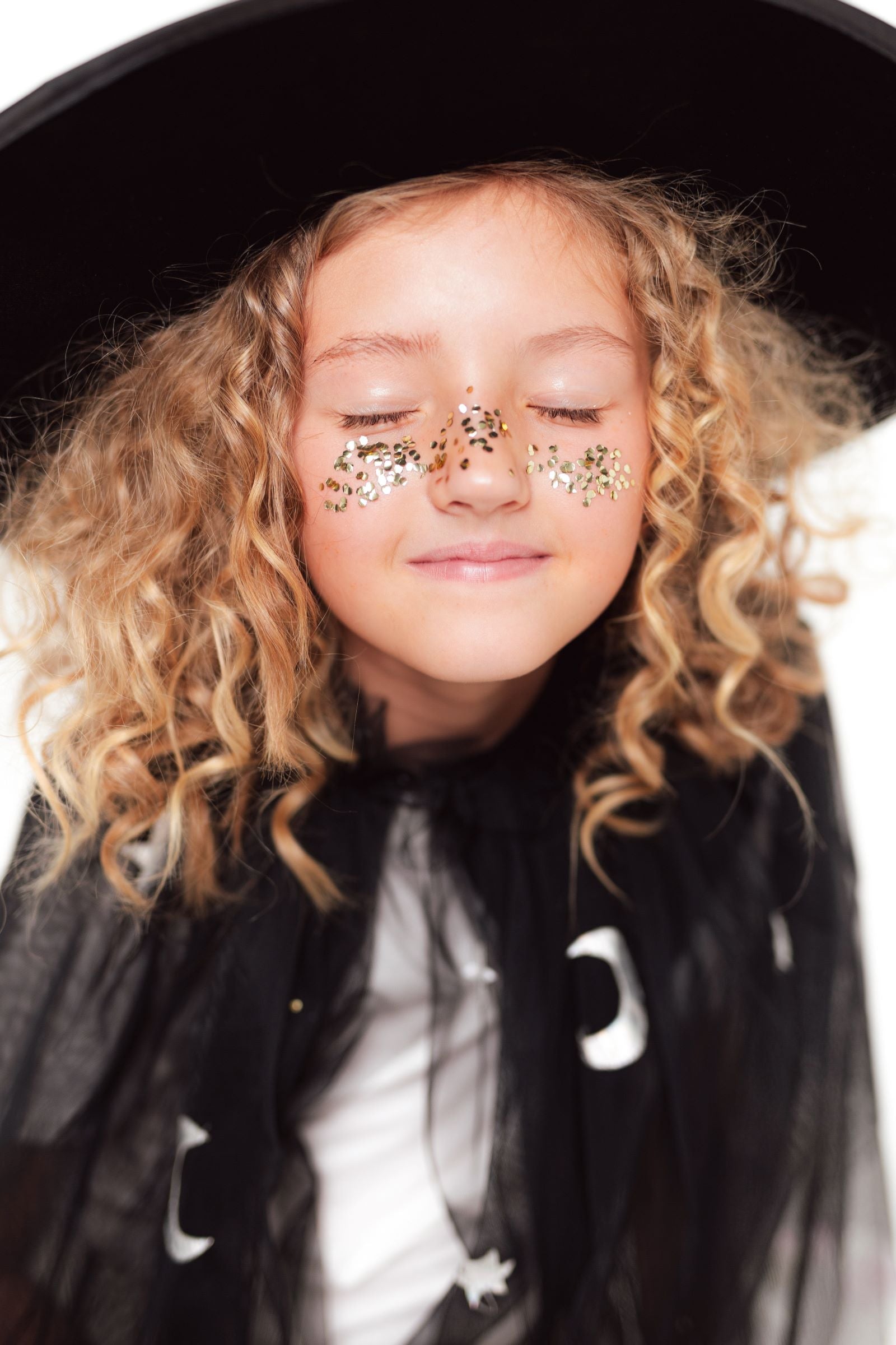 A close-up of a child's face with eyes closed, with gold glitter applied across the bridge of the nose and cheeks to create a freckle effect, and wearing a black witch hat.
