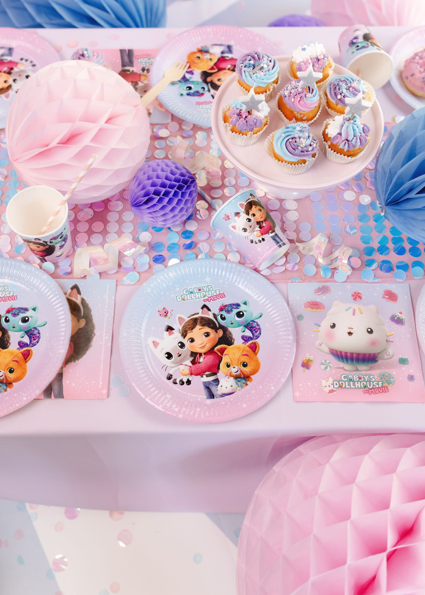 Gabby's Dollhouse paper napkins arranged on a pink party table next to matching character plates and cupcakes.