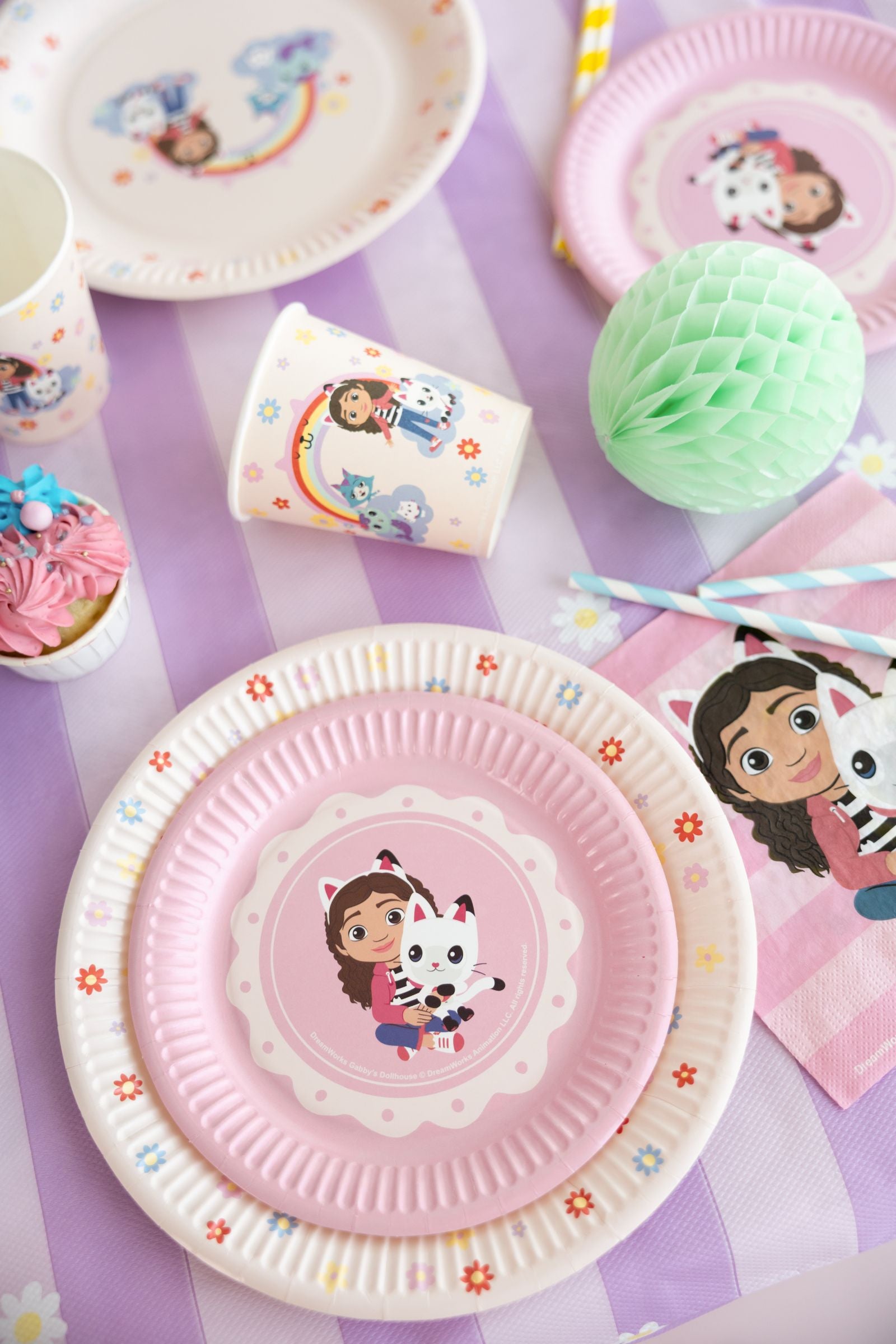 Children's Gabby's Dollhouse party table setting with paper plates, cups, and napkins featuring a cartoon character on a purple tablecloth.