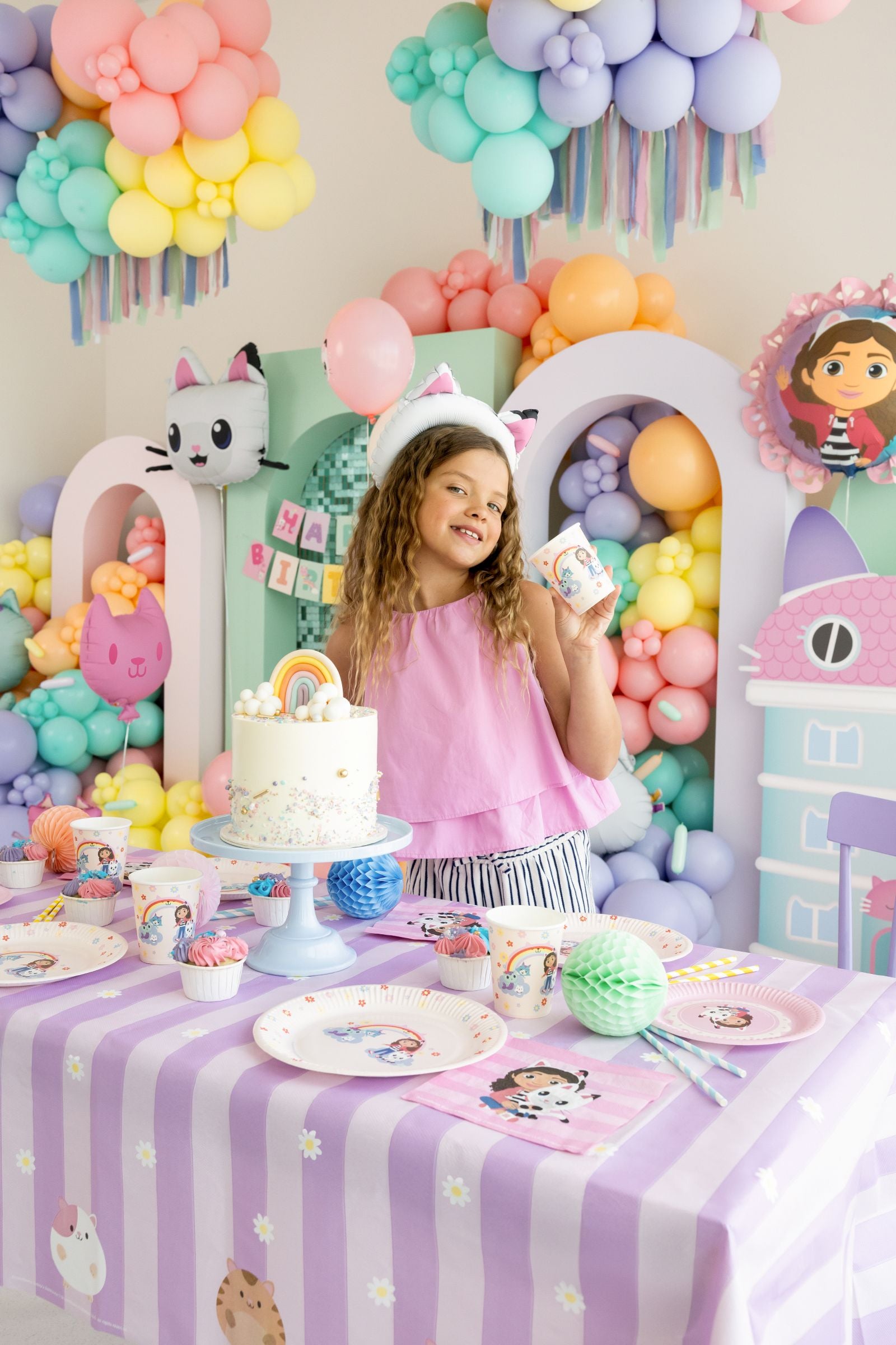 Child at a Gabby's Dollhouse themed birthday party with colorful balloons and decorations, including a cake and tableware.