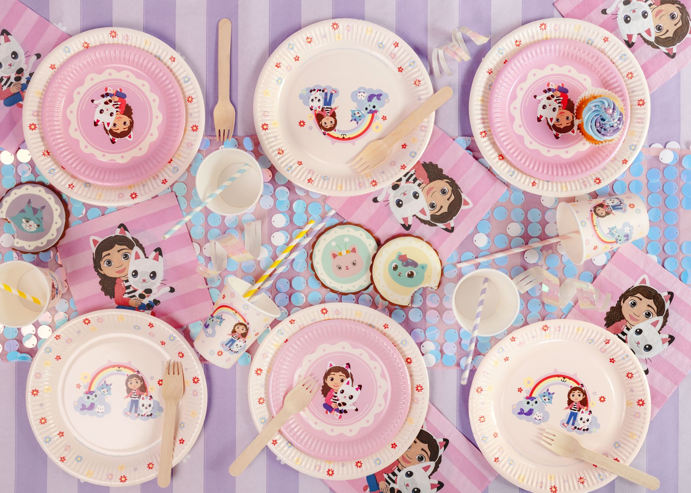 Children's party tableware with Gabby's Dollhouse cartoon character plates, cups, and cutlery on a purple and pink background.