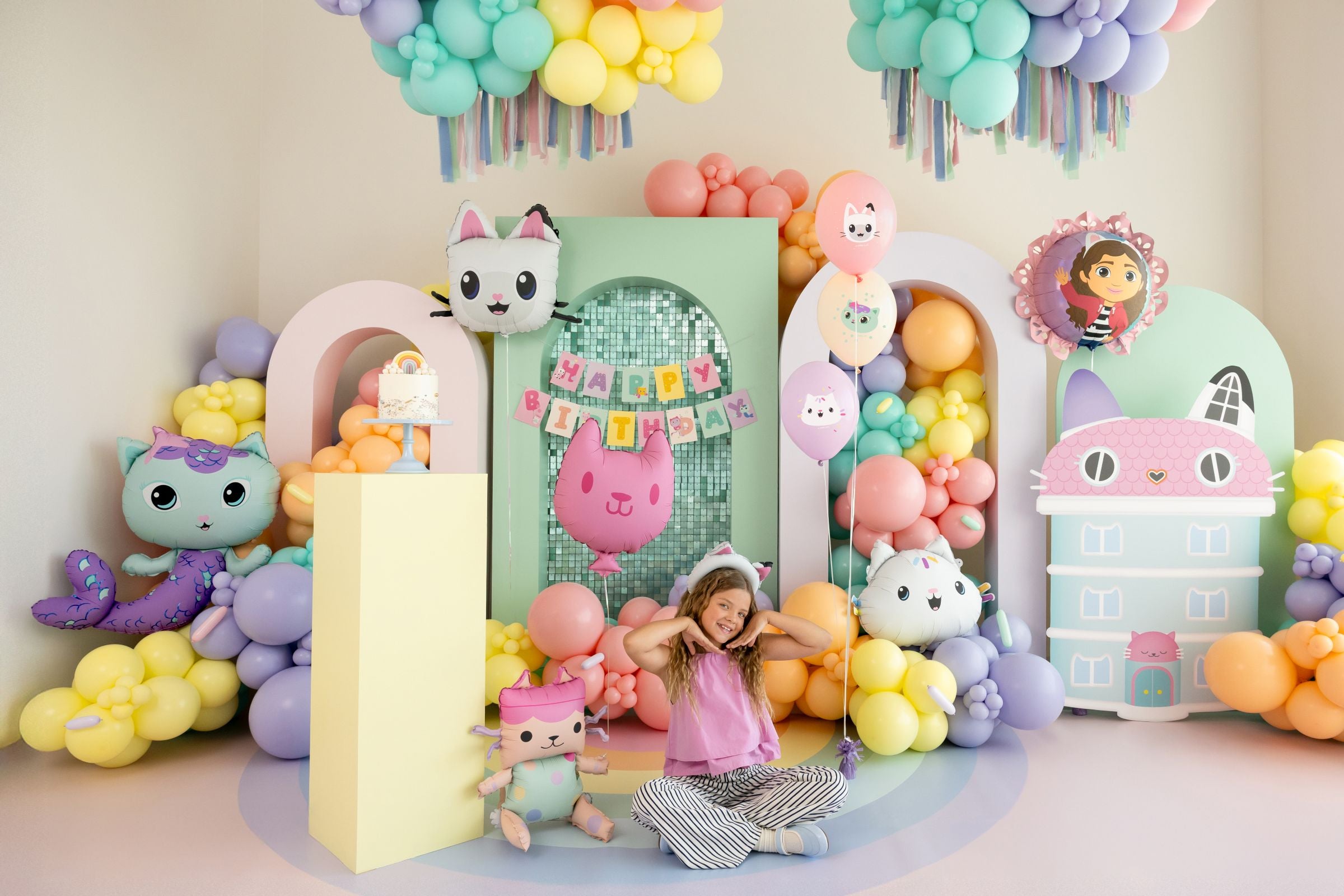A young girl sitting on the floor surrounded by Gabby's Dollhouse themed party decorations and character balloons.