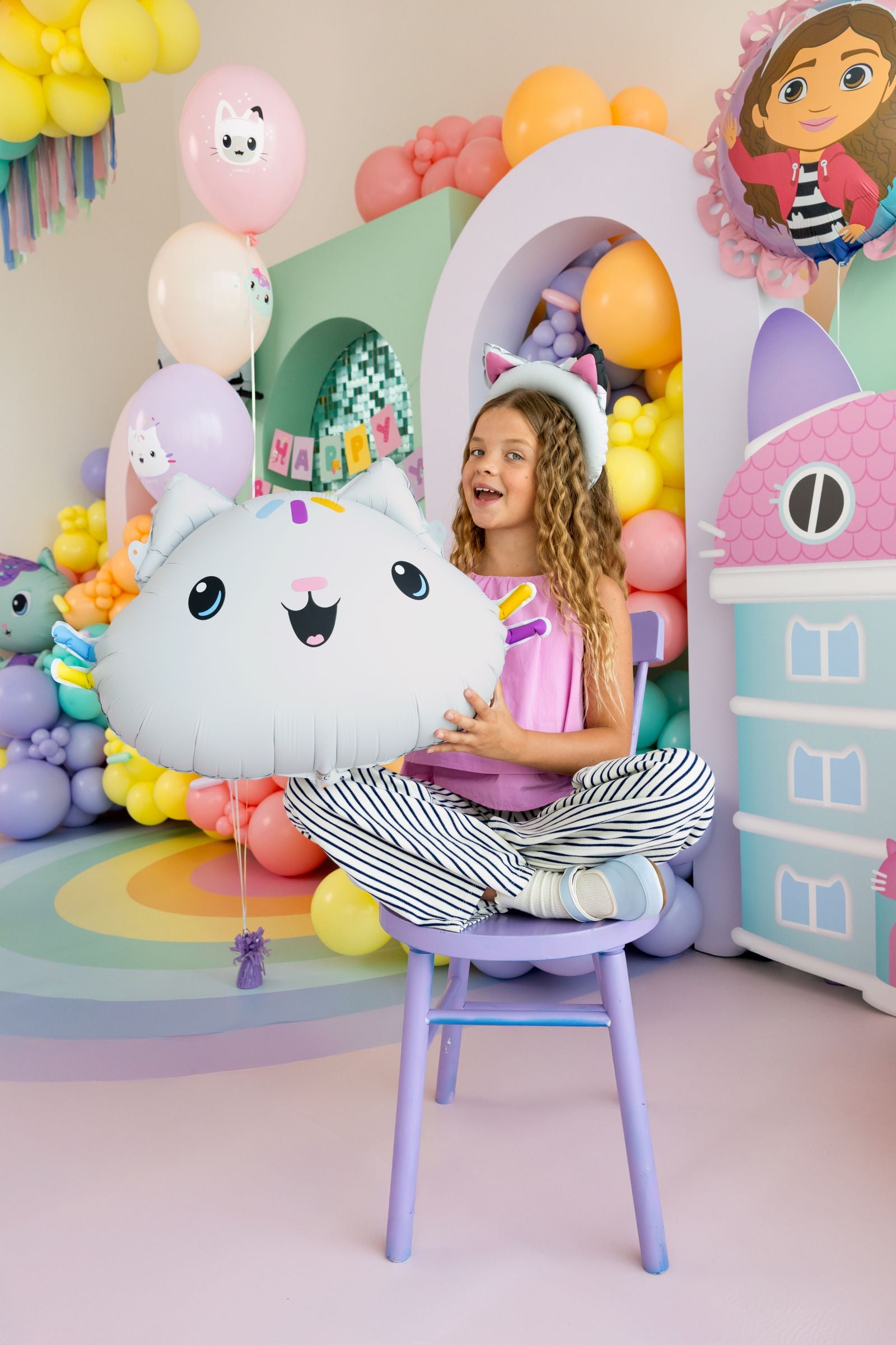 Child sitting on a chair holding a large Cakey Balloon in a colorful, playful Gabby's Dollhouse celebration featuring a Cakey foil balloon, colorful balloon arches, and matching table settings.