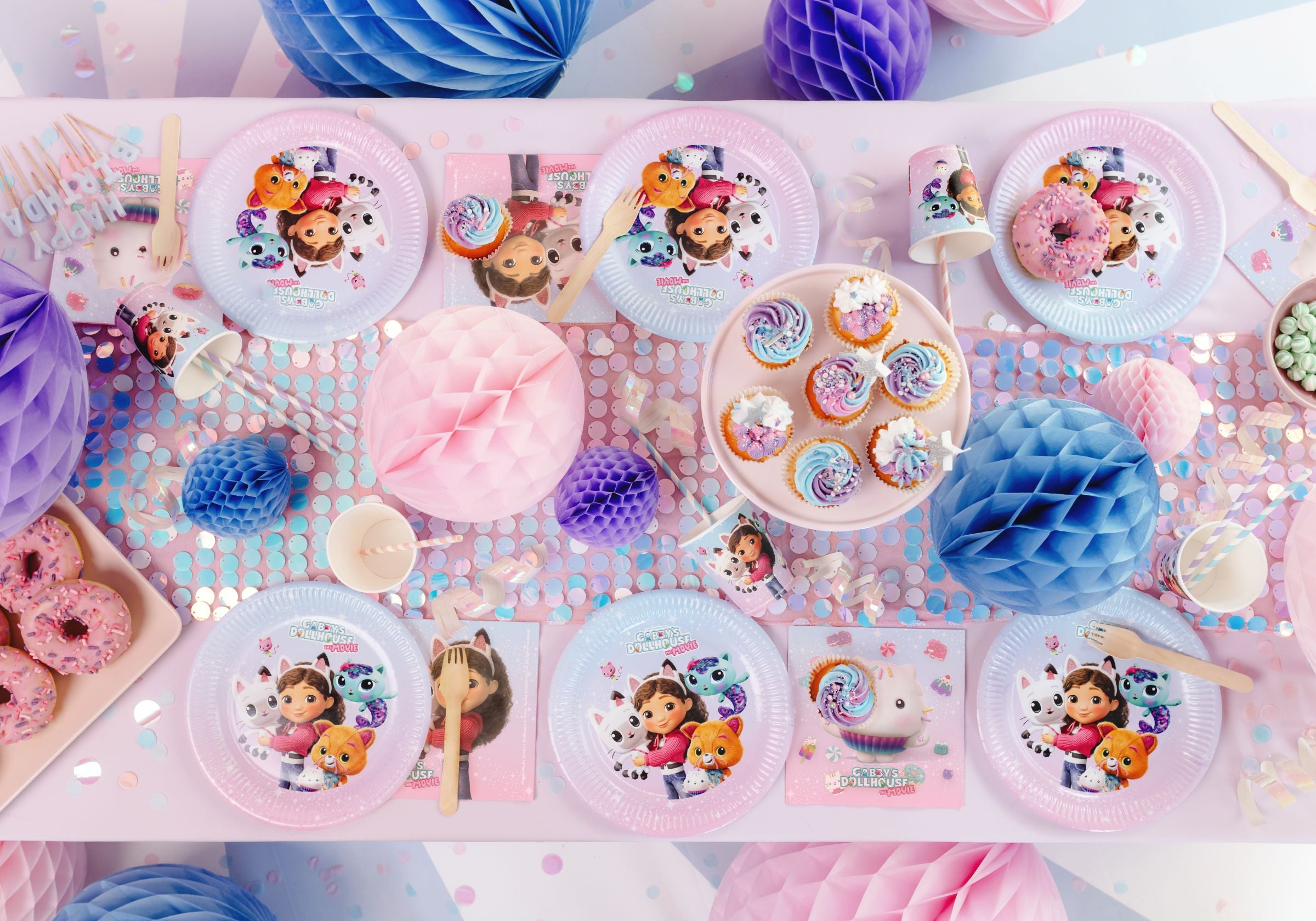 A top-down view of a beautifully styled party table featuring Gabby's Dollhouse The Movie 23cm paper plates, matching cups, napkins, pink frosted donuts, and cupcakes arranged on a pastel sequin table runner.