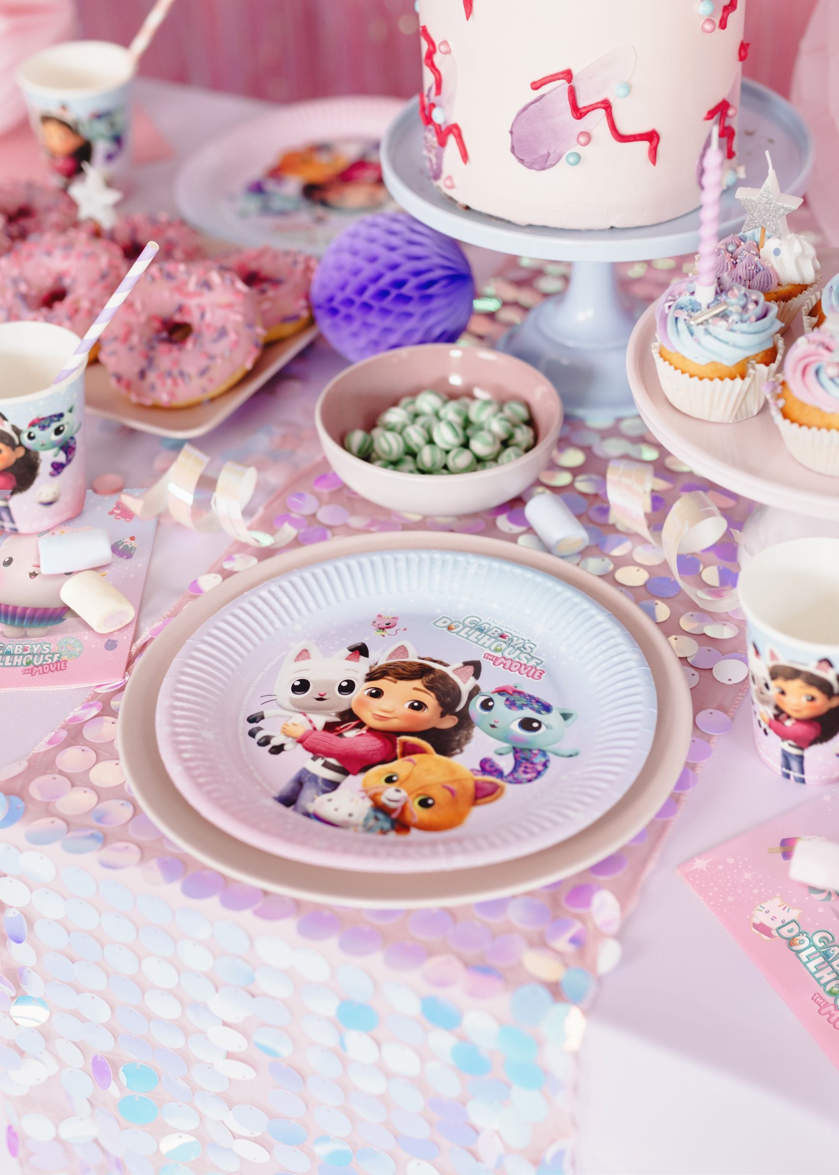 A close-up of a Gabby's Dollhouse The Movie paper plate resting on a shimmering pink sequin table runner, surrounded by matching cups, napkins, mint candies, and beautifully decorated cupcakes.