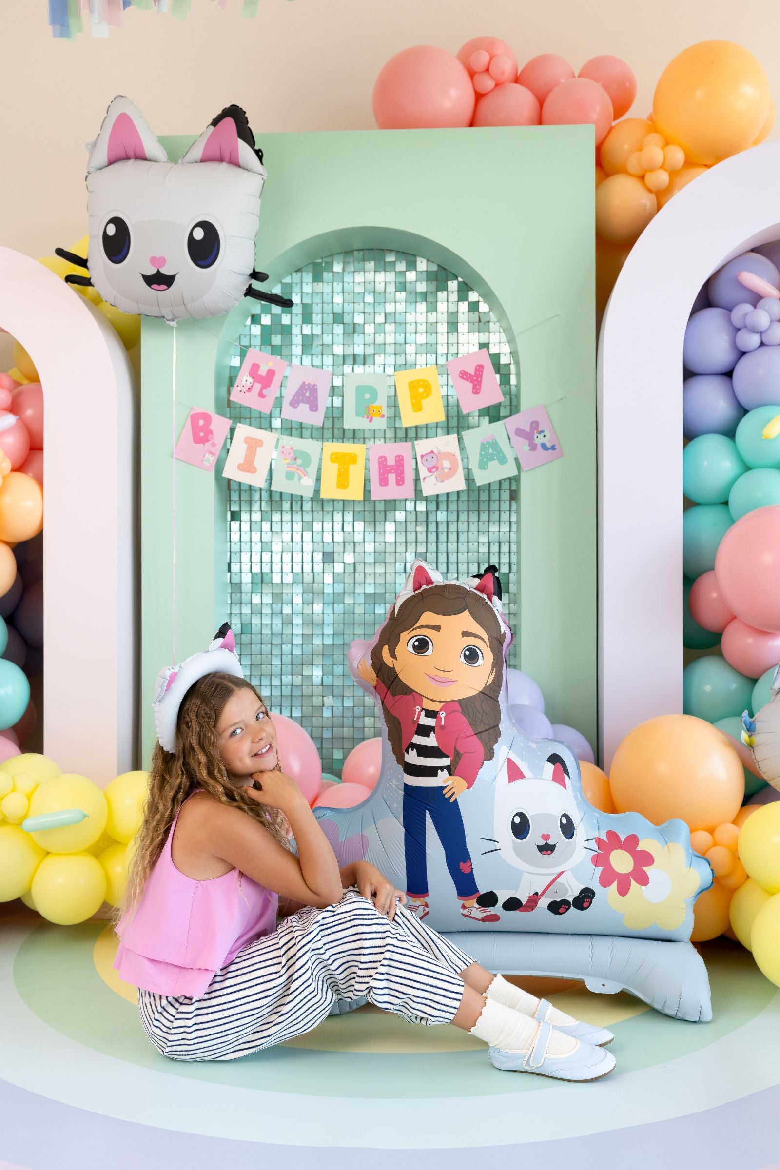 A young girl sitting next to a large standing Gabby’s Dollhouse character balloon in a festive party room.