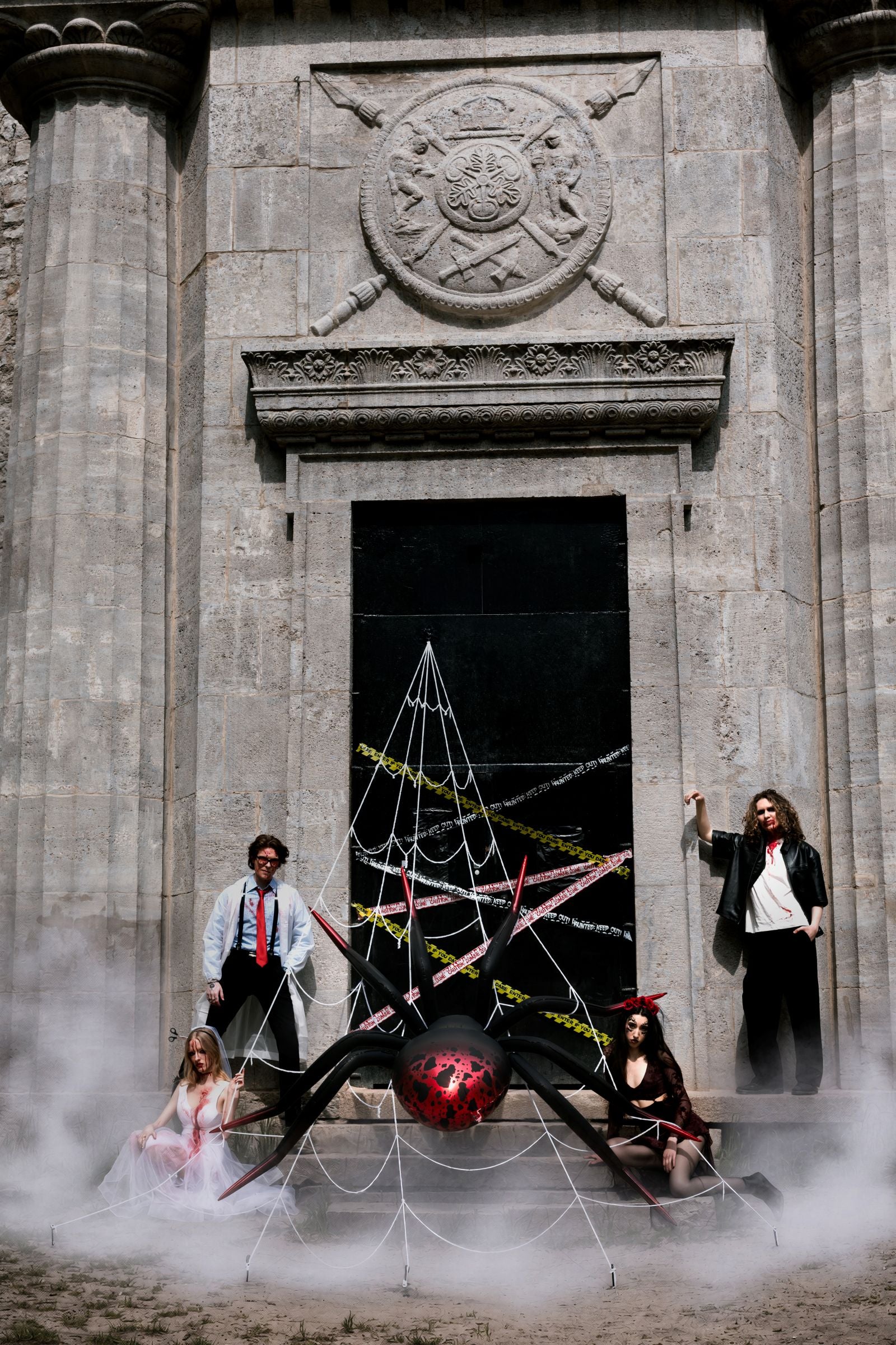 A group of four people posing in front of a gothic stone building doorway, surrounding a large black spider decoration with white webbing and a red spider ball on the floor, with fog rising around them.