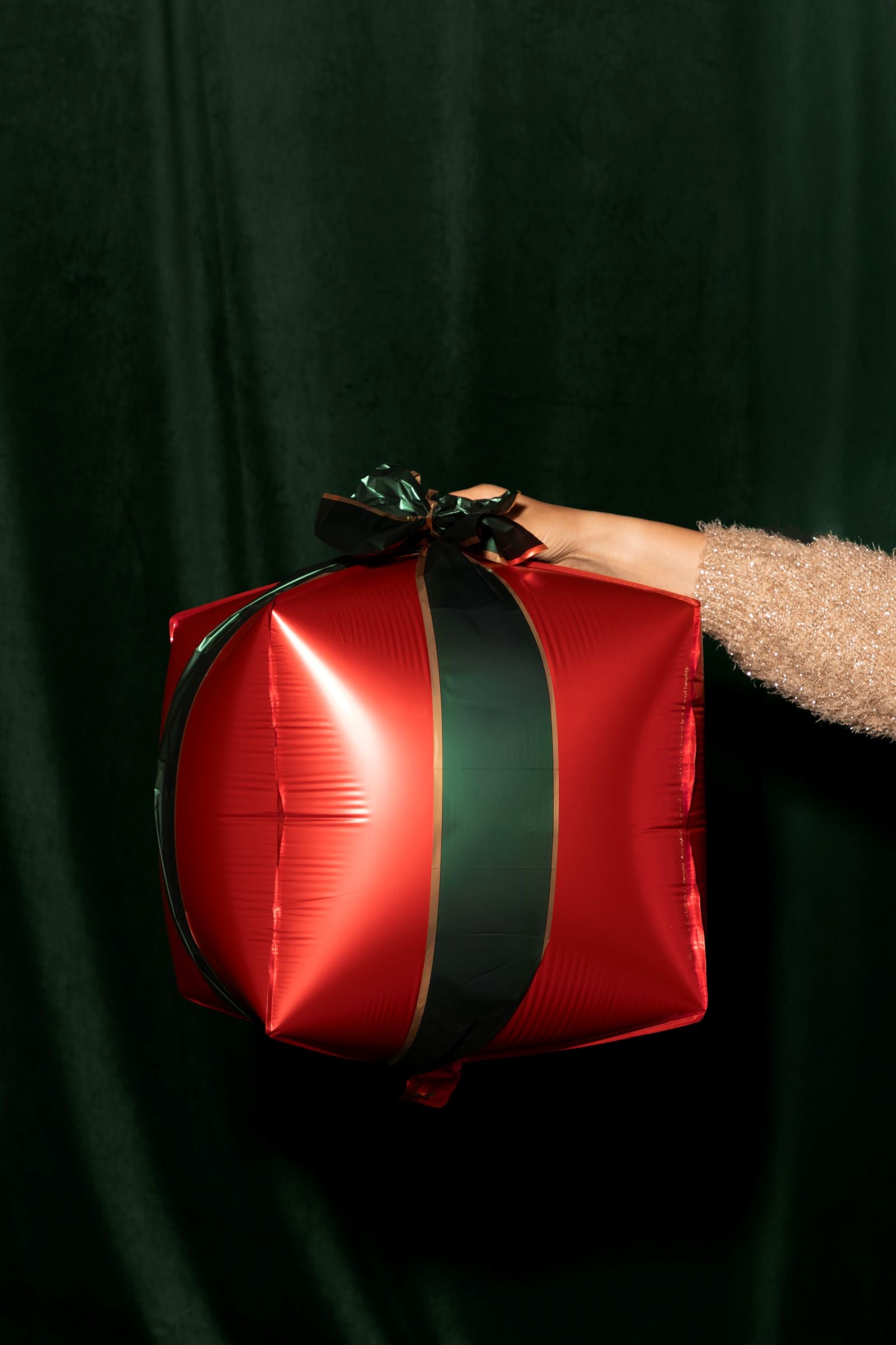 A person's hand holding a red and green foil balloon shaped like a Christmas present against a dark green curtain.