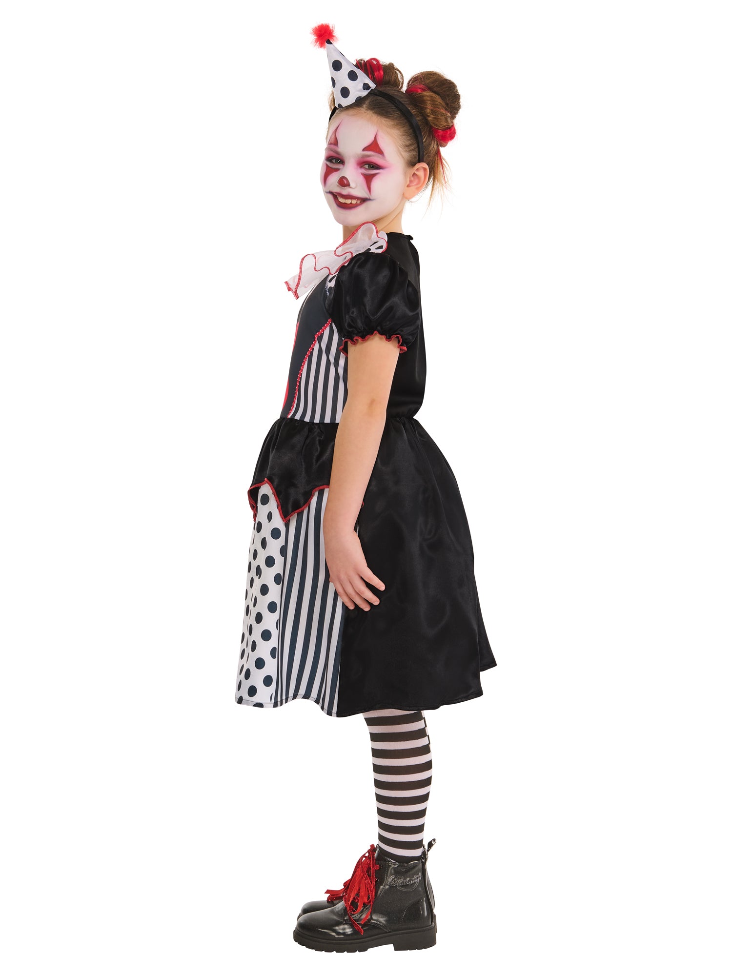 A side profile of a child in a black and white clown costume, striking a pose. The costume includes a dress with mixed polka dot and striped patterns, a ruffled collar, and red buttons, worn with striped leggings and a headband with a small hat, against a white background.