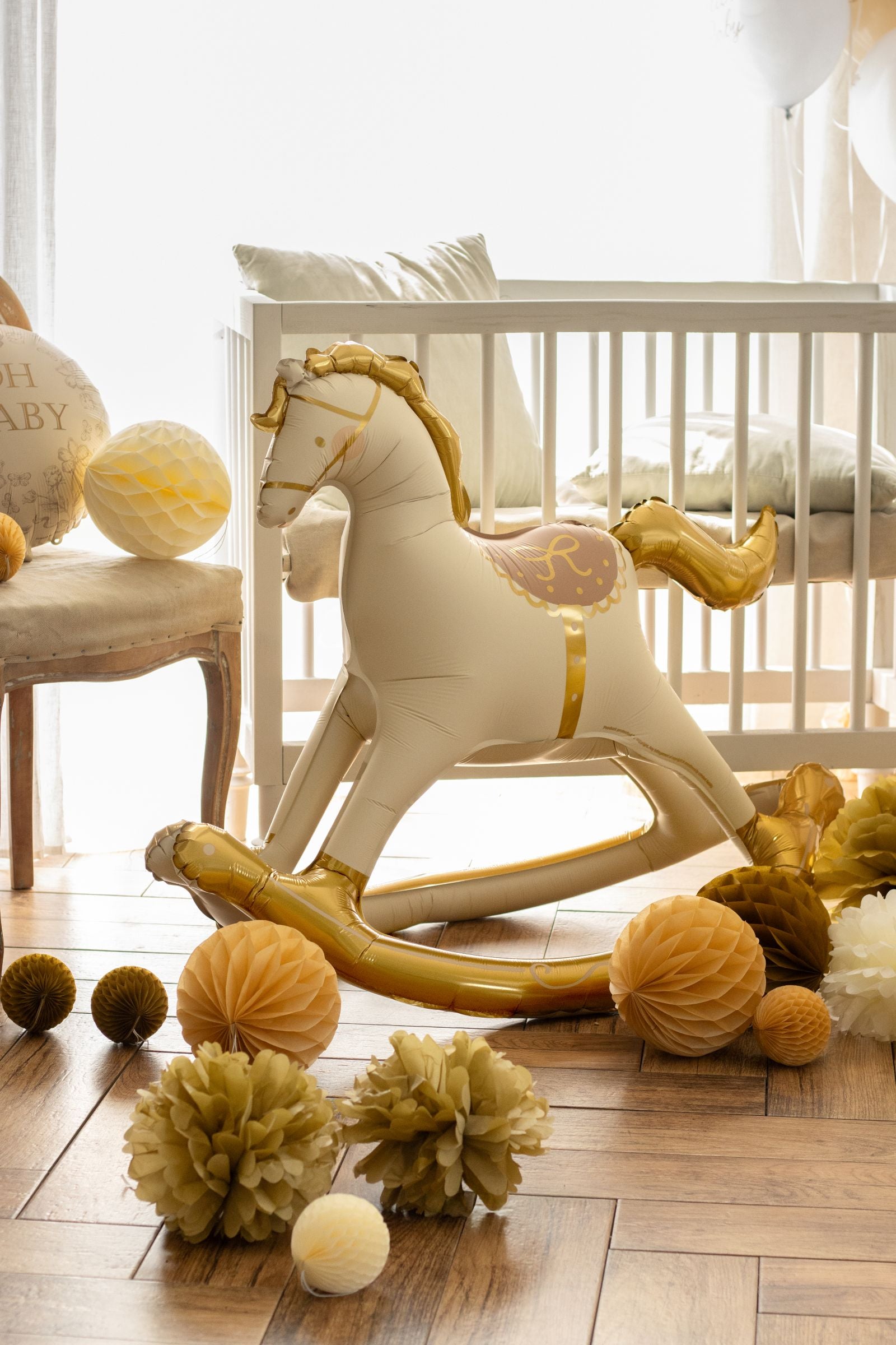 A gold rocking horse foil balloon standing next to a white baby crib in a decorated nursery.