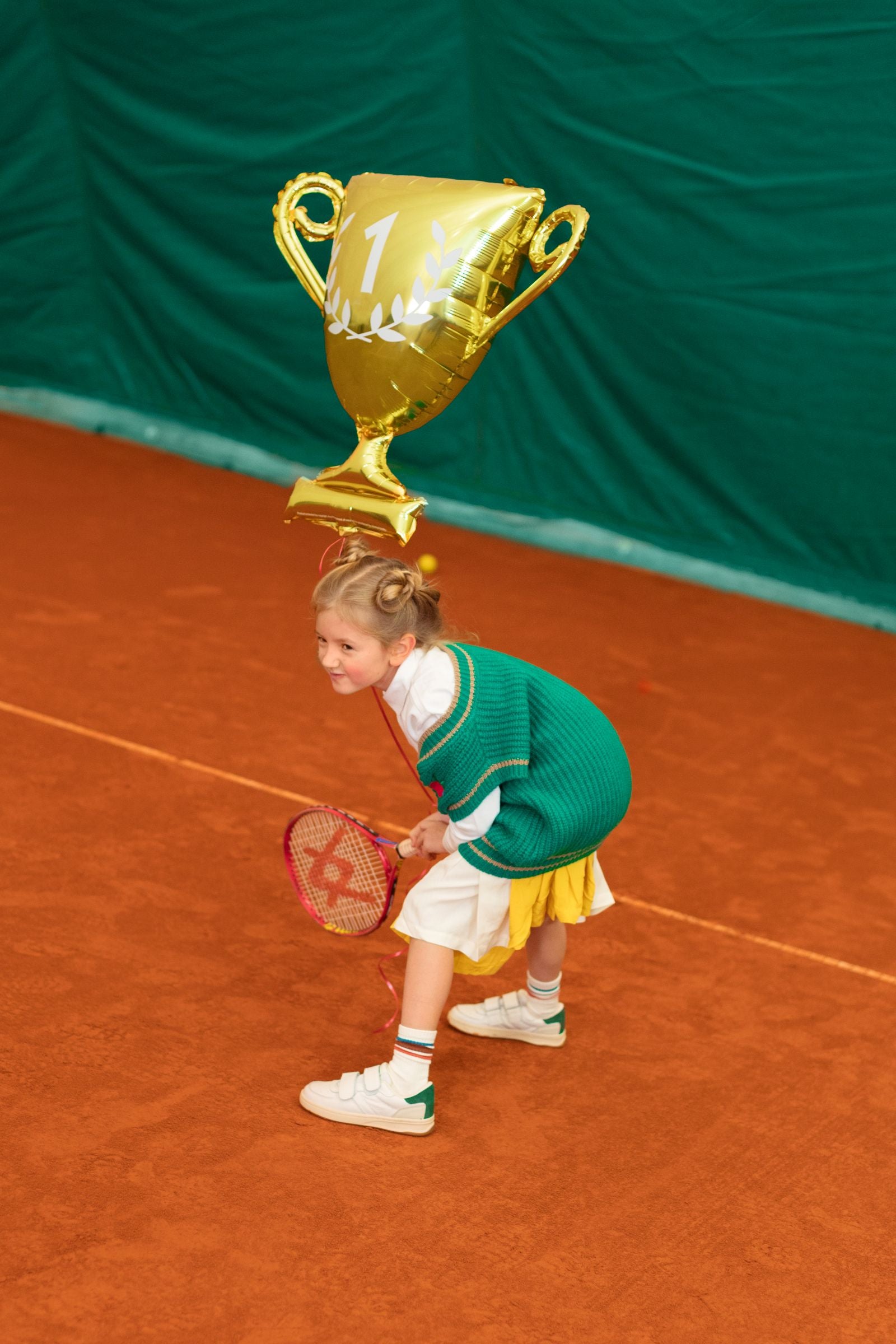 Young girl on a tennis court with a large gold trophy balloon above her head.