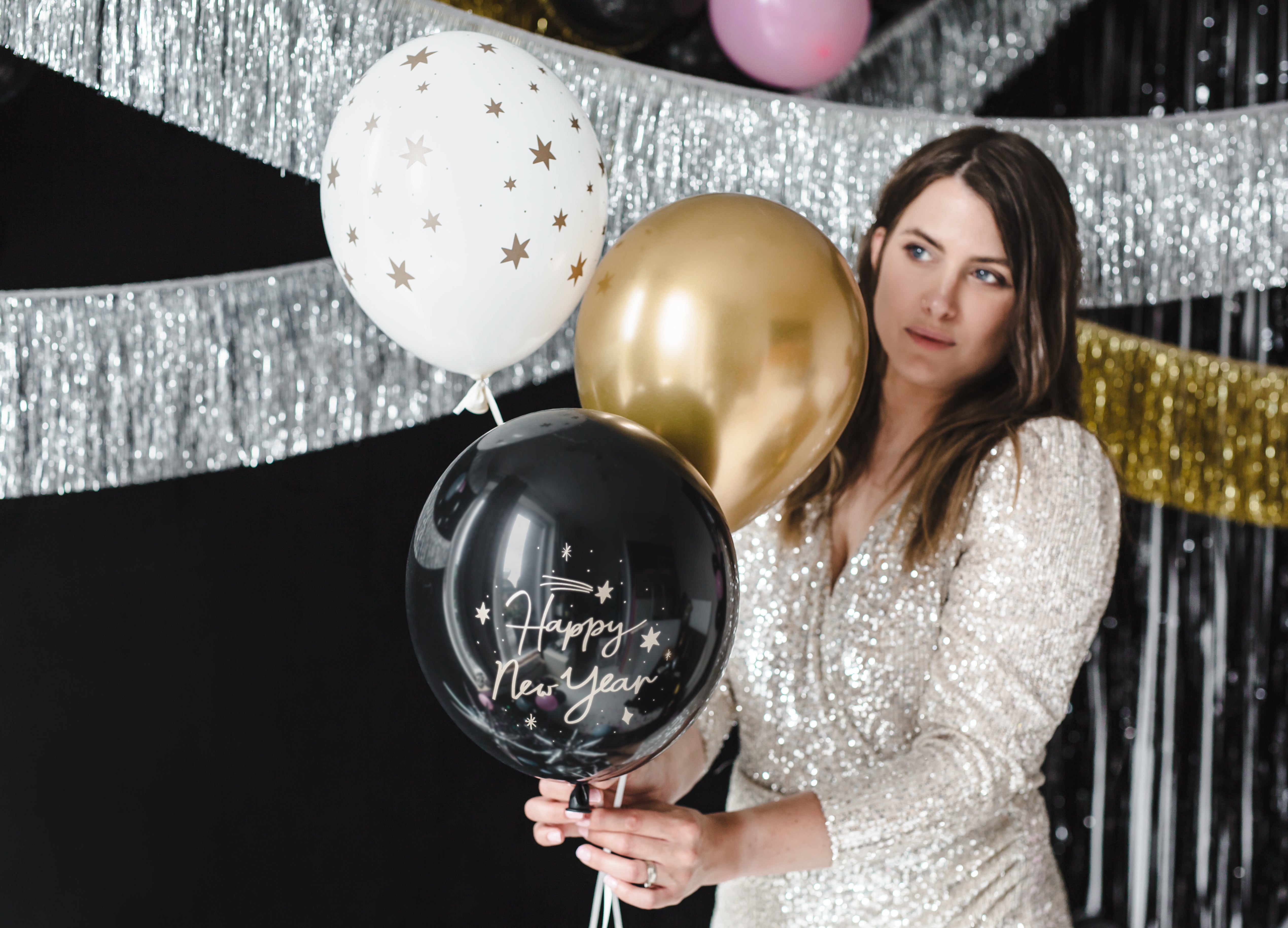 A woman in a silver sequined dress holding a bunch of black, white, and gold Happy New Year balloons against a festive tinsel backdrop.