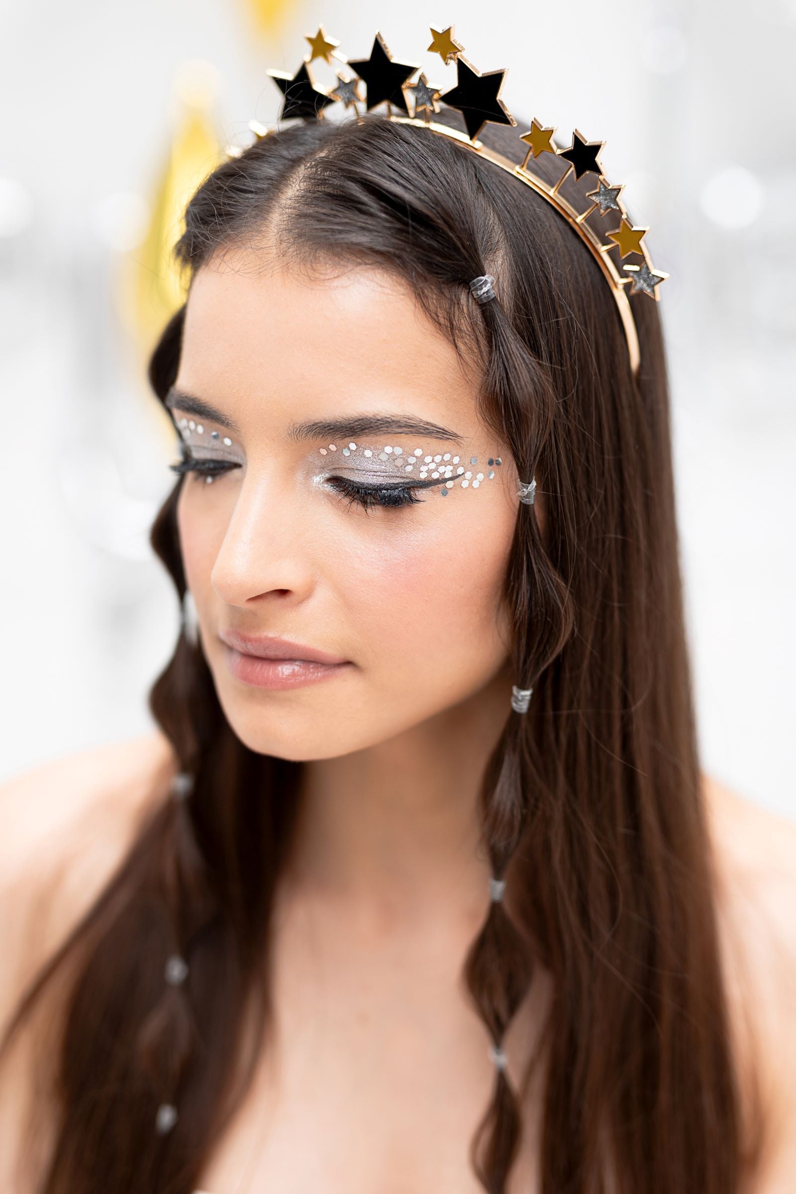 A person with a decorative headband and braids, with their eyes closed, wearing holographic glitter applied in a freckle pattern across the bridge of the nose and cheeks, against a light background.