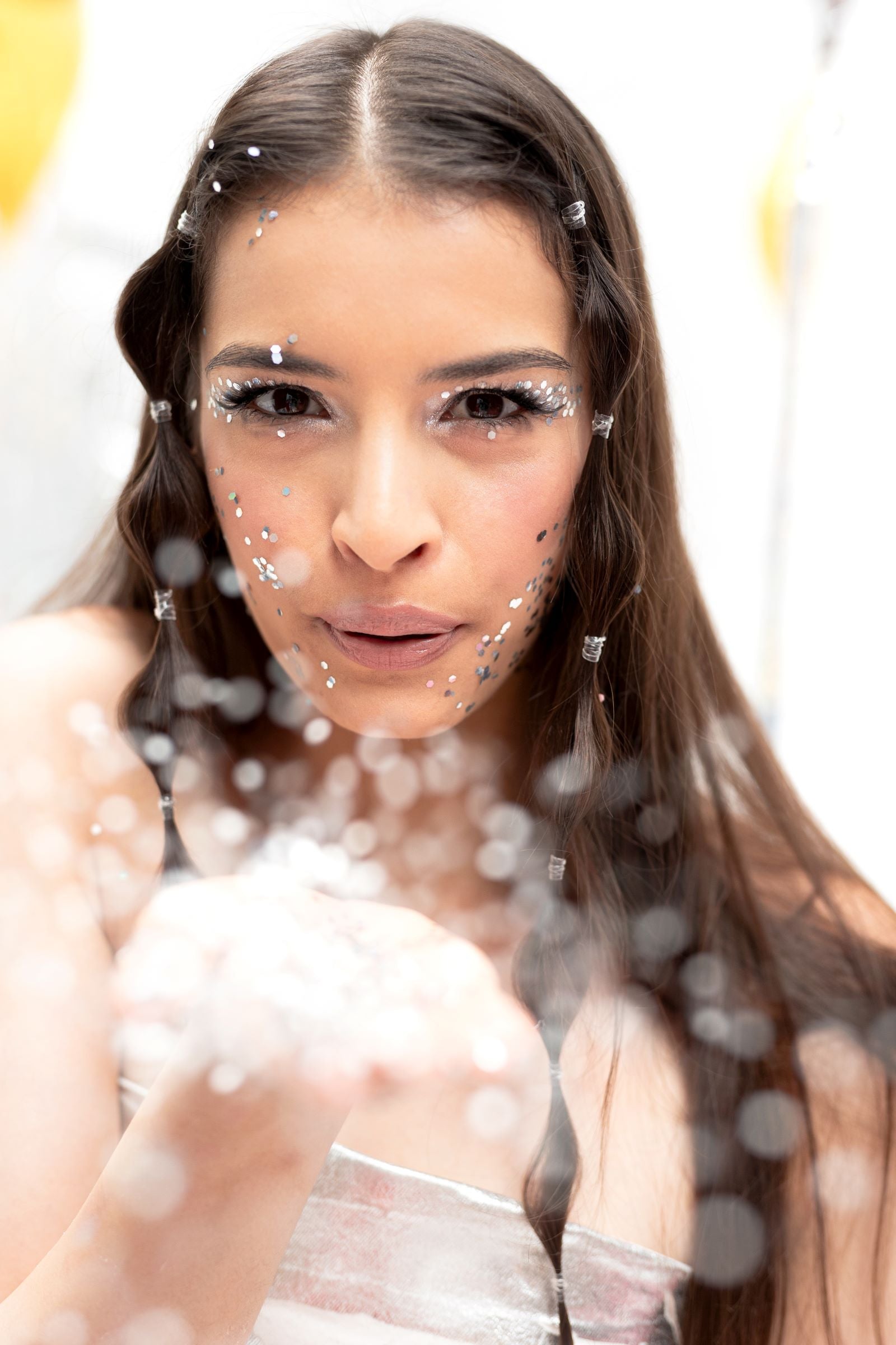 A close-up of a person blowing a handful of silver holographic glitter towards the camera. Glitter is also applied across the bridge of the person's nose and cheeks.