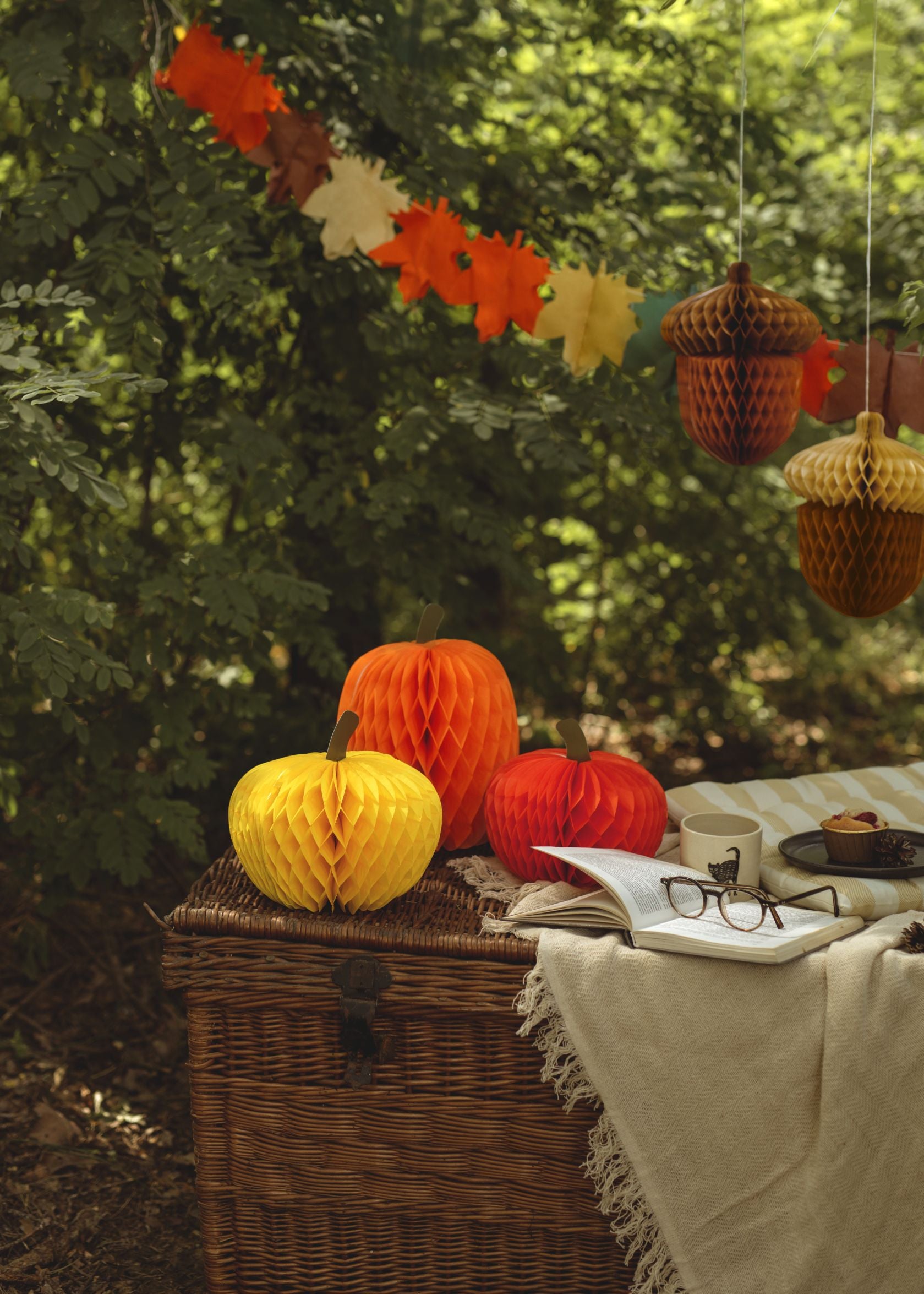 An outdoor autumn scene featuring three honeycomb paper pumpkins (orange, yellow, red) of varying sizes placed on a woven basket. In the background, an autumn leaf garland hangs, along with two hanging honeycomb acorn decorations, amidst lush green foliage.