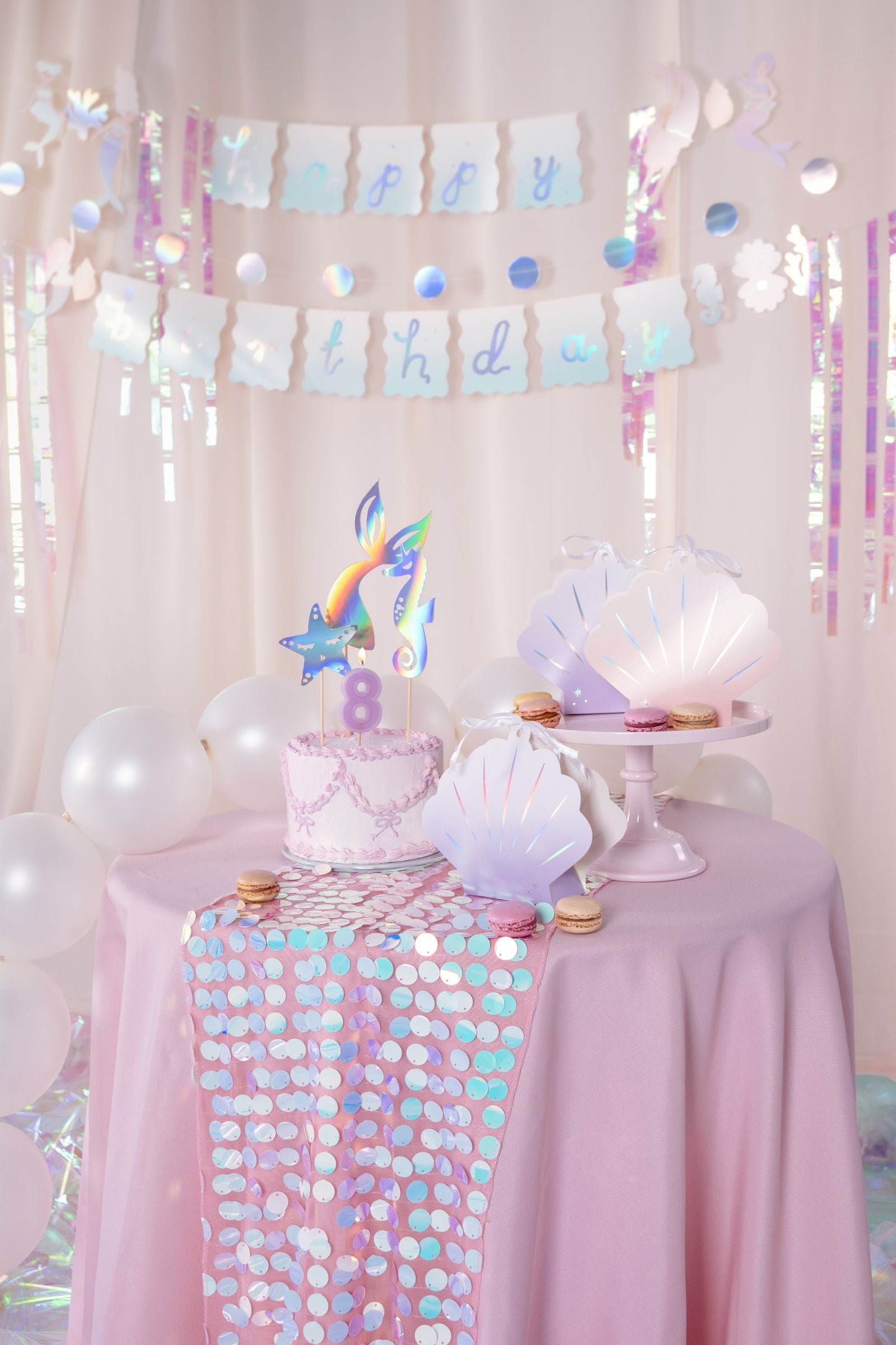 A festive mermaid-themed dessert table with a light pink tablecloth. An iridescent sequin table runner is draped diagonally across the table, showcasing its shimmering, multi-colored sequins, alongside a birthday cake, seashell-shaped snack boxes, macarons, and a "Happy Birthday" banner in the background.