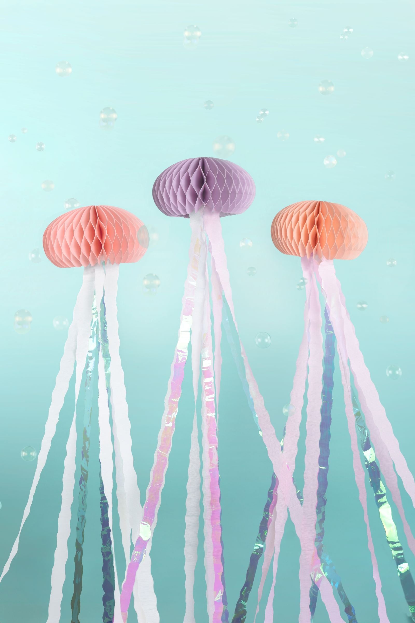 Three colorful tissue paper jellyfish decorations hanging vertically against a light blue background with bubbles. The jellyfish have honeycomb tops in orange, purple, and peach, with long, shimmering iridescent and white ribbon streamers acting as tentacles.