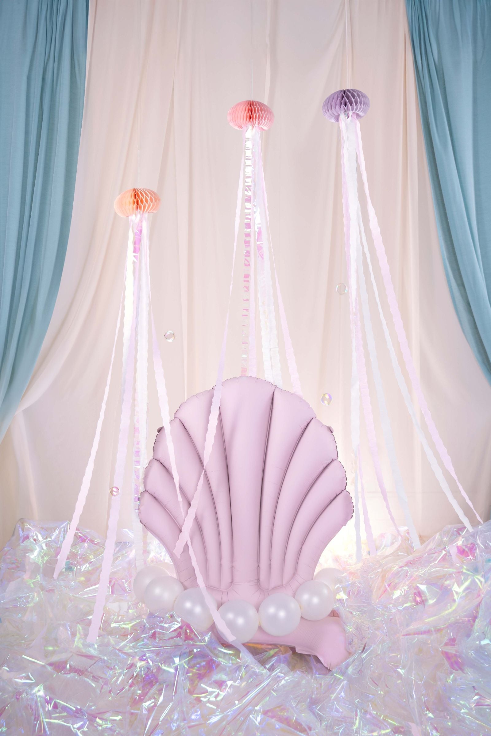 A whimsical mermaid-themed party backdrop featuring a pink seashell-shaped chair with pearl-like balloons, set on an iridescent floor. Above the chair, three colorful jellyfish tissue paper decorations with long streamers hang from the ceiling, against a backdrop of draped blue and white curtains.