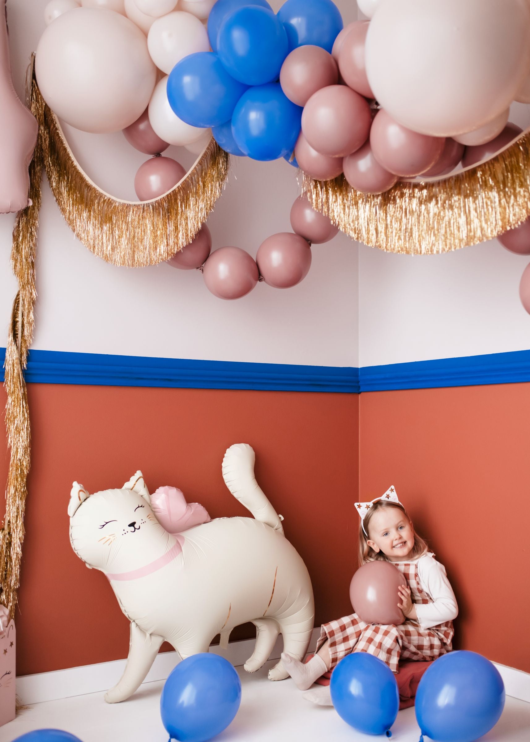 A happy child sitting next to a large standing white cat foil balloon in a decorated party room.