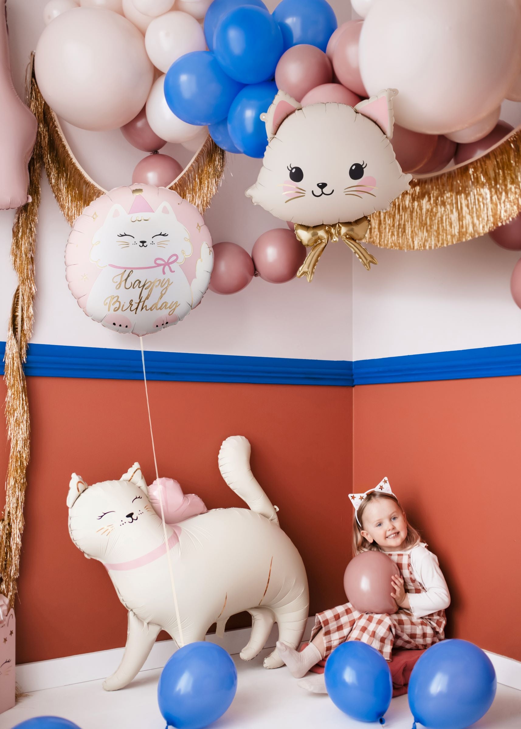 A child celebrating with a variety of cat-shaped and round kitty foil balloons amidst a balloon arch.