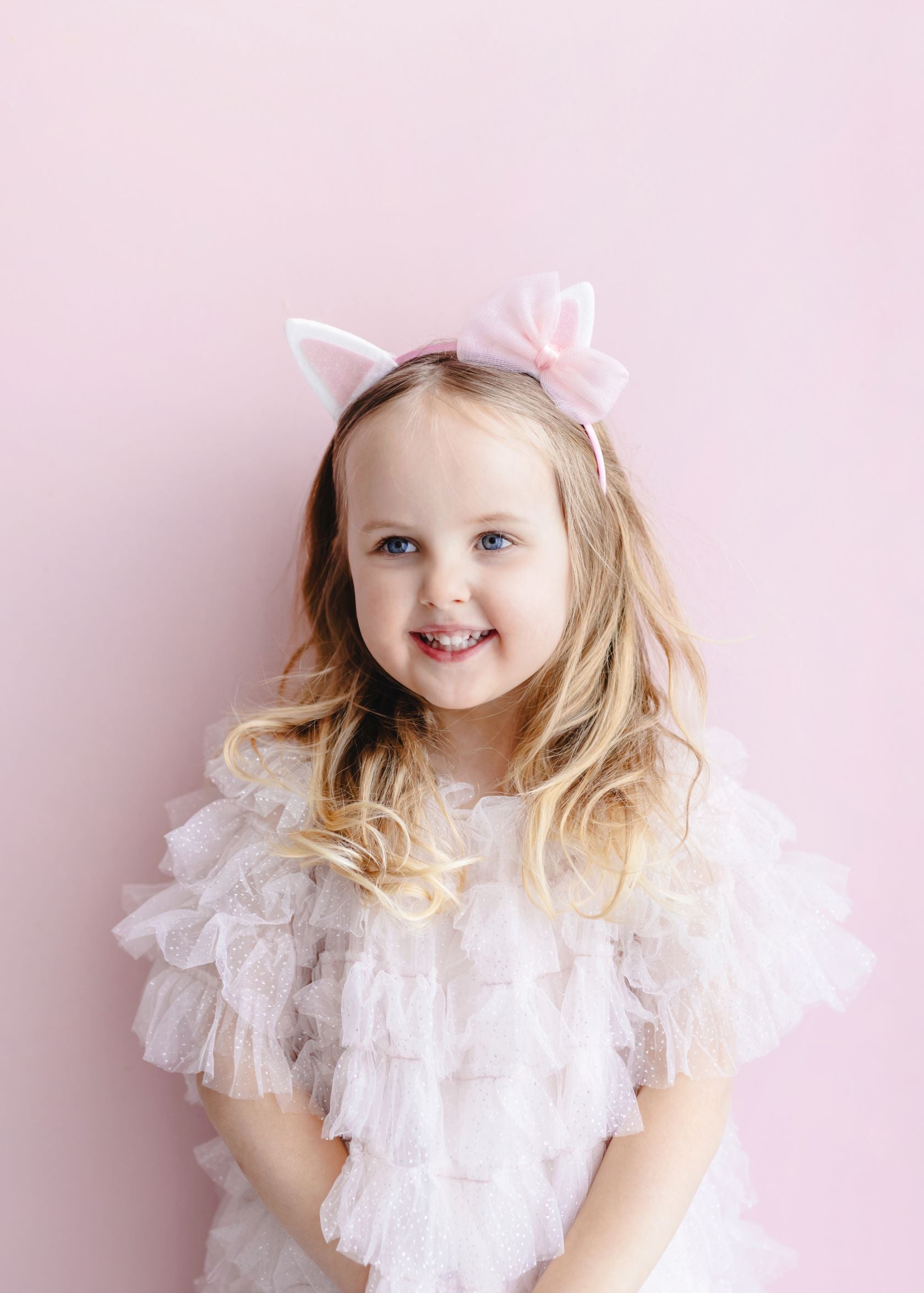A young child smiling, wearing a pink dress and a pink headband with white cat ears and a pink organza bow, against a light pink background.