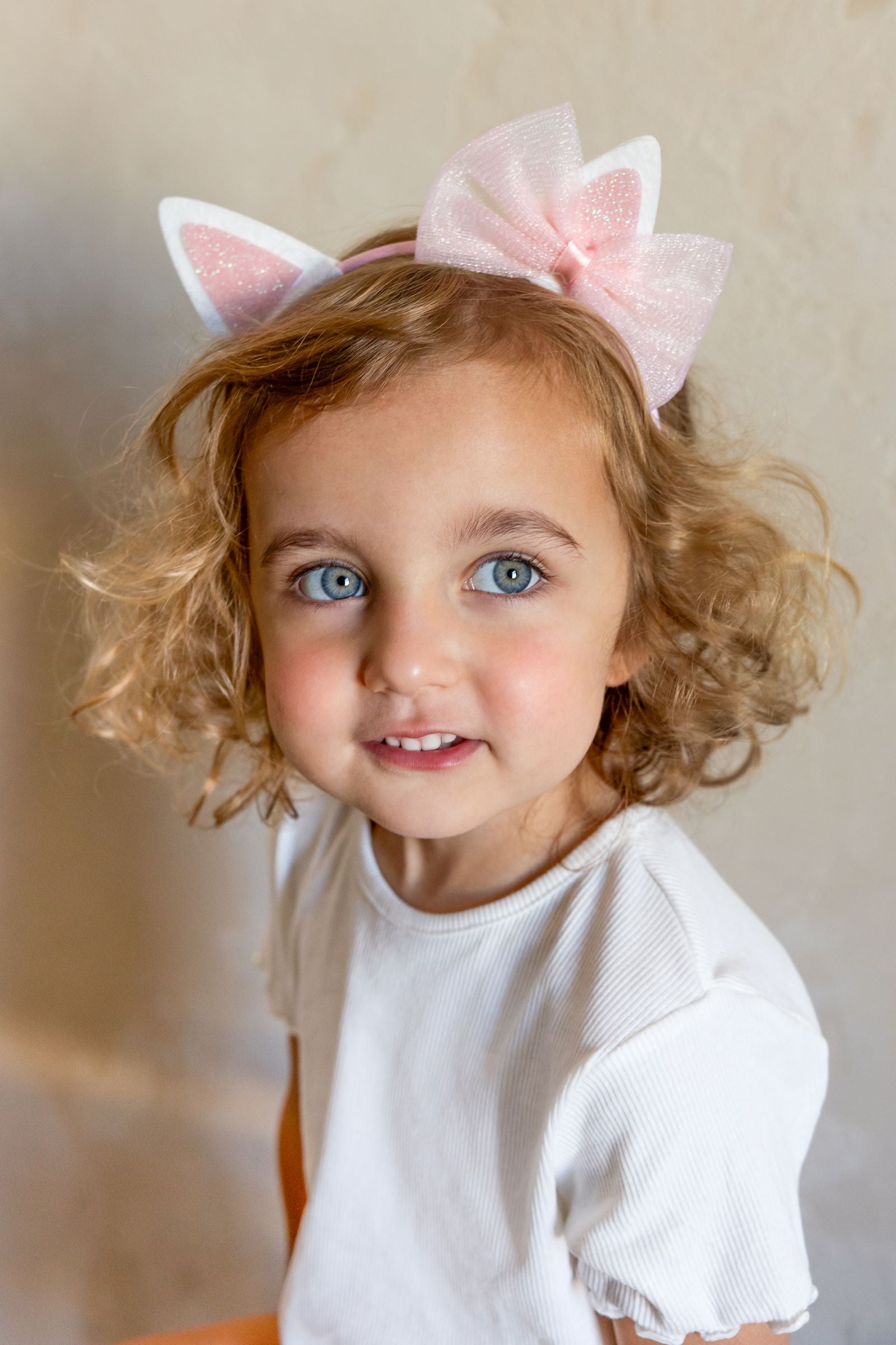 A young child smiling, wearing a white t-shirt and a pink headband with white cat ears and a pink organza bow, against a textured beige background.